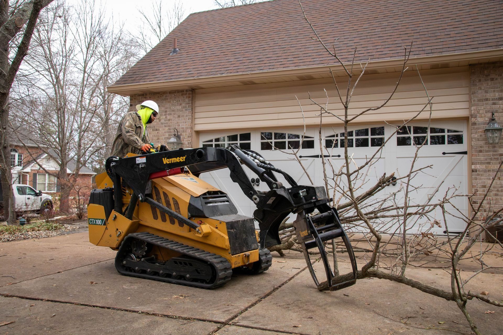 A worker uses a compact track loader with a grapple attachment to remove tree branches near a garage.