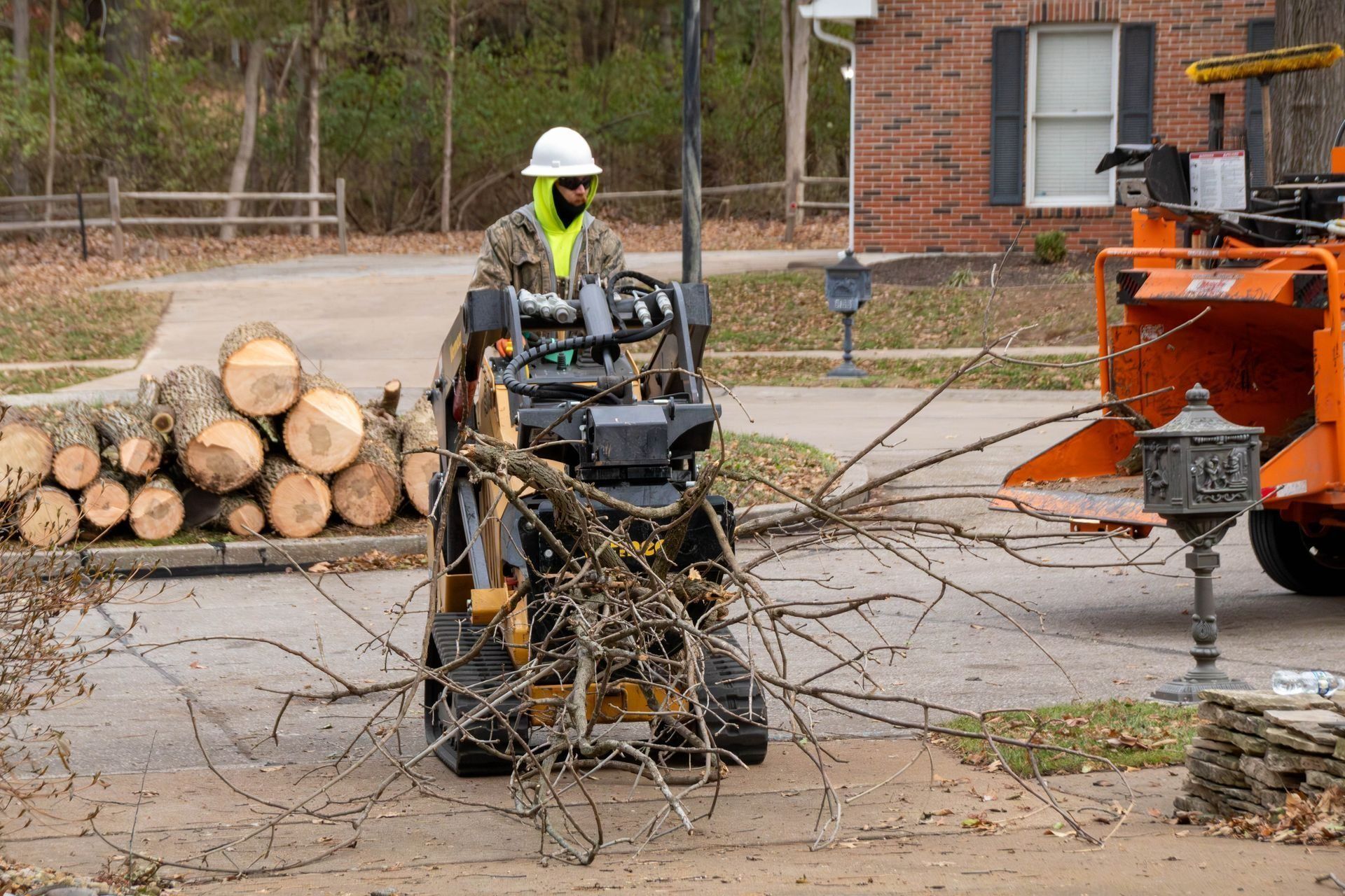 Worker in a white hard hat operates a compact loader to move tree branches near a wood chipper, driveway, and stacked logs.