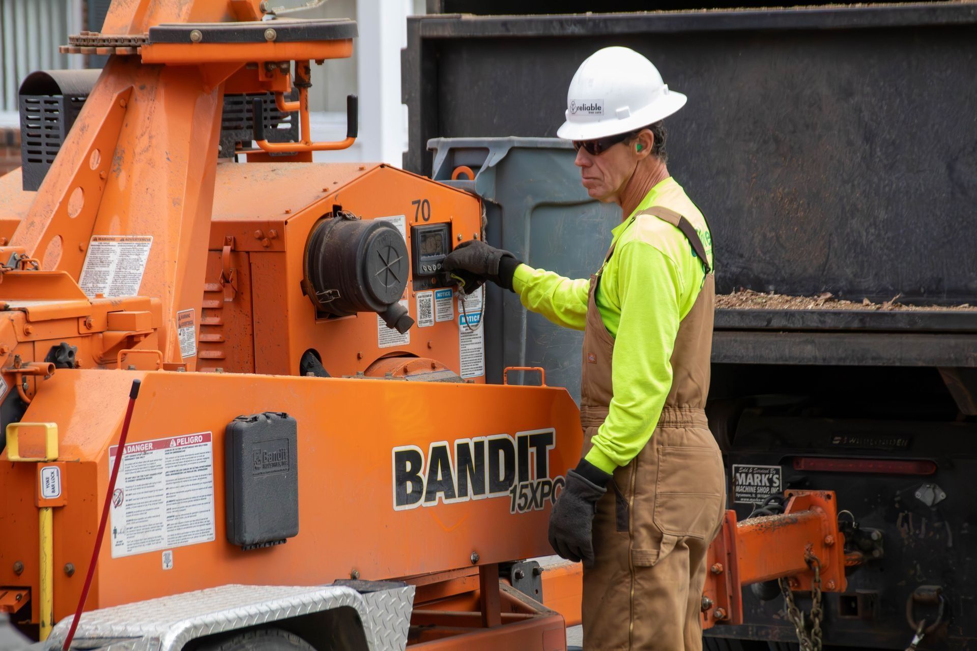 Man operating an orange Bandit wood chipper, wearing a hard hat, overalls, and safety glasses.
