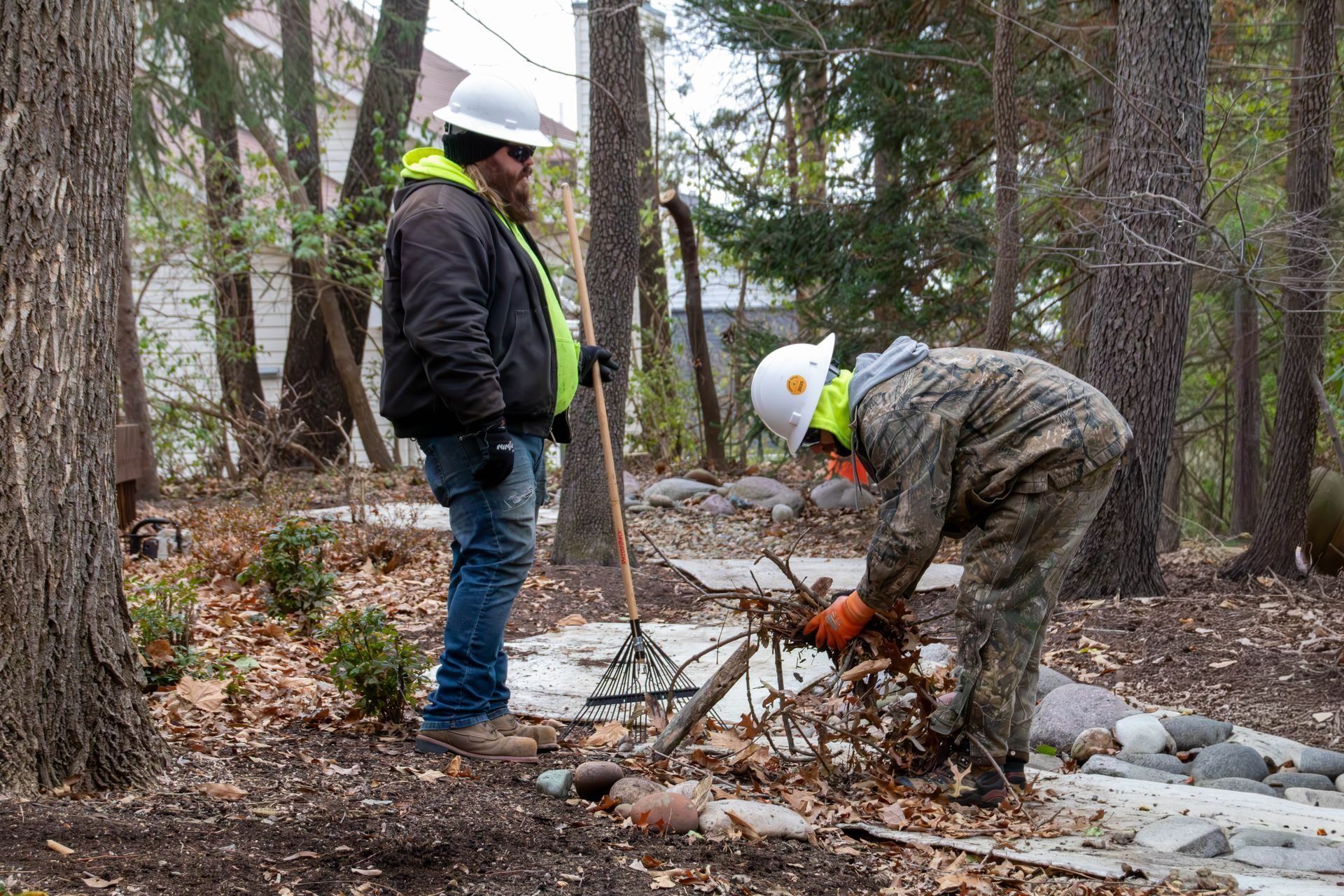 Two people raking leaves outdoors. One observes, the other leans over a pile. Trees surround them.