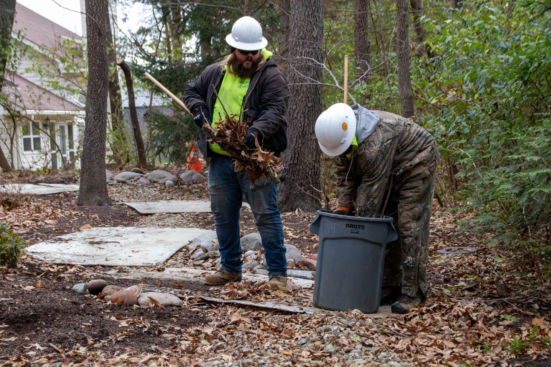 Two workers in hard hats rake and collect leaves in a wooded area, filling a trash can.