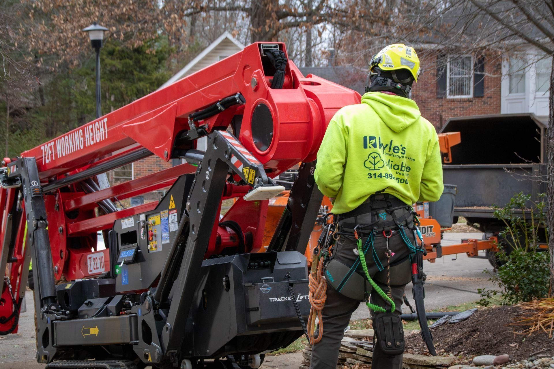 Arborist in a yellow hoodie operating an aerial lift to trim a tree in front of a house.