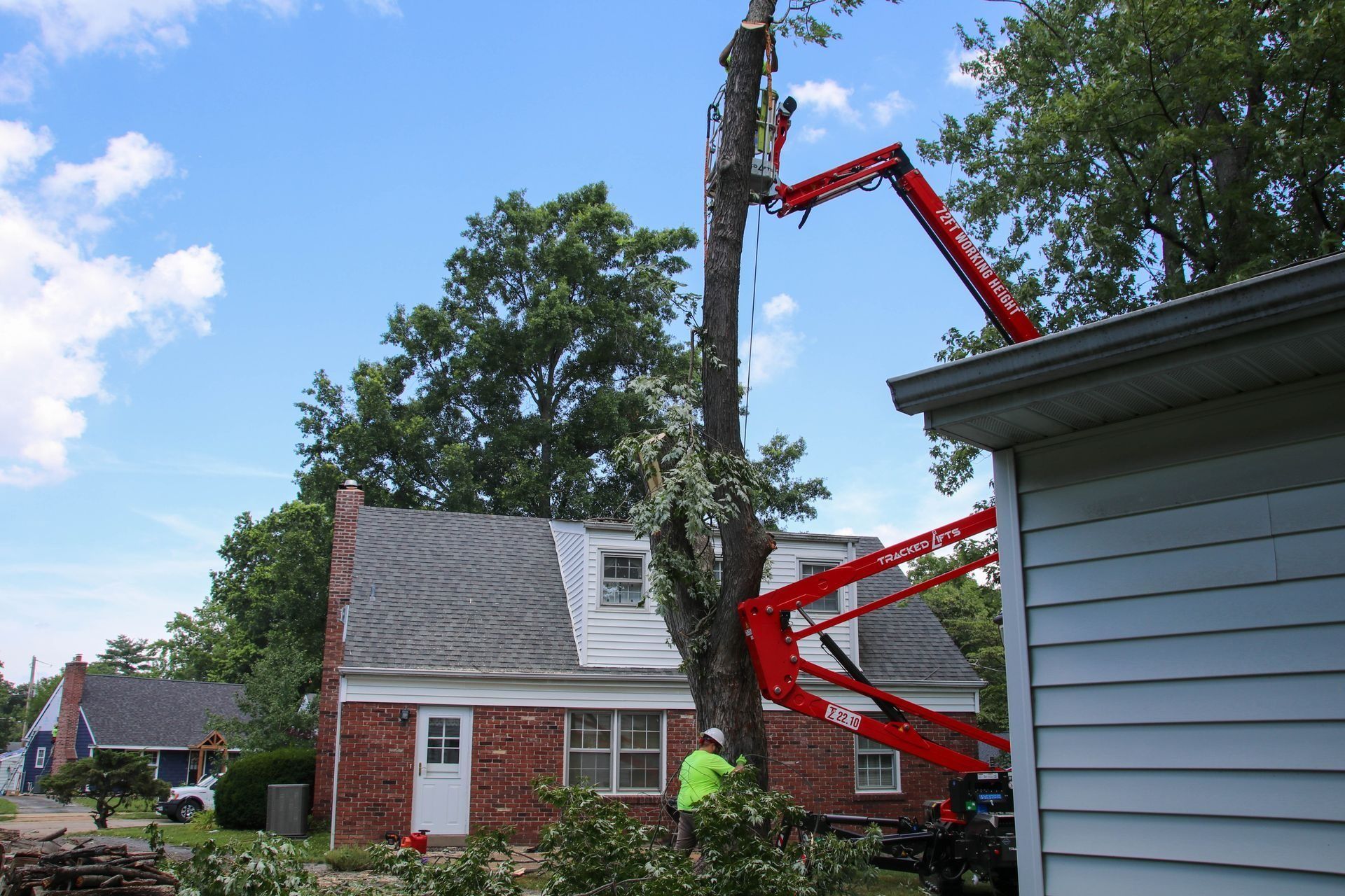A tree being trimmed next to a house by workers using a red boom lift on a sunny day.