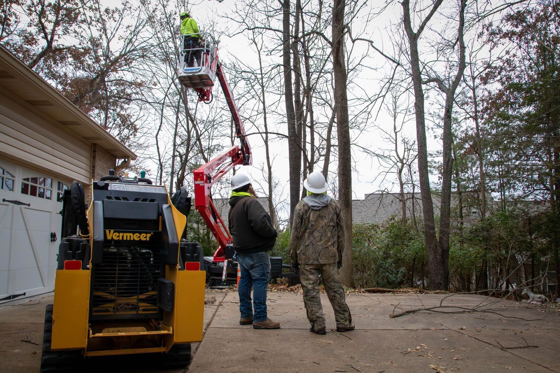 Tree trimming crew using a lift near a house. Yellow skid steer, two workers observing the worker in the lift.