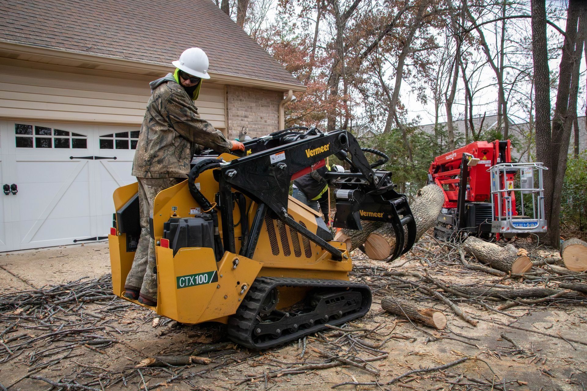 A person operates a yellow skid steer with a tree shear. Logs are in the yard, a house and tree lift in the background.