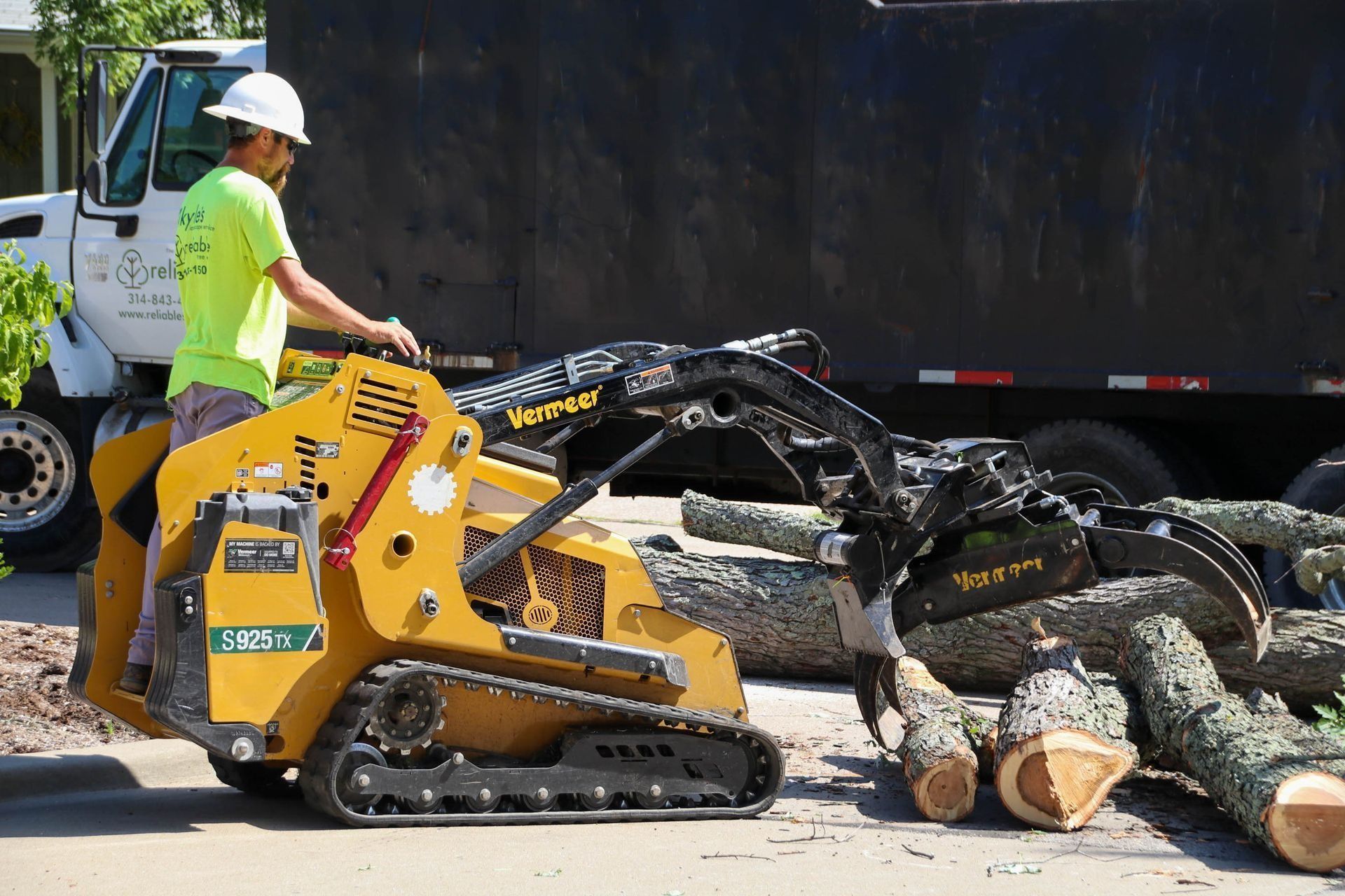 Man operating a yellow skid steer with grapple, loading cut logs onto a trailer in a residential setting.