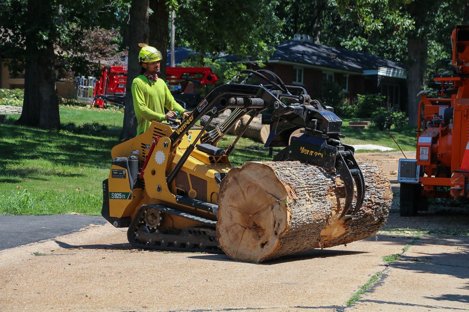 A person in a safety vest operates a yellow mini-skid steer to lift a large tree trunk on a driveway.