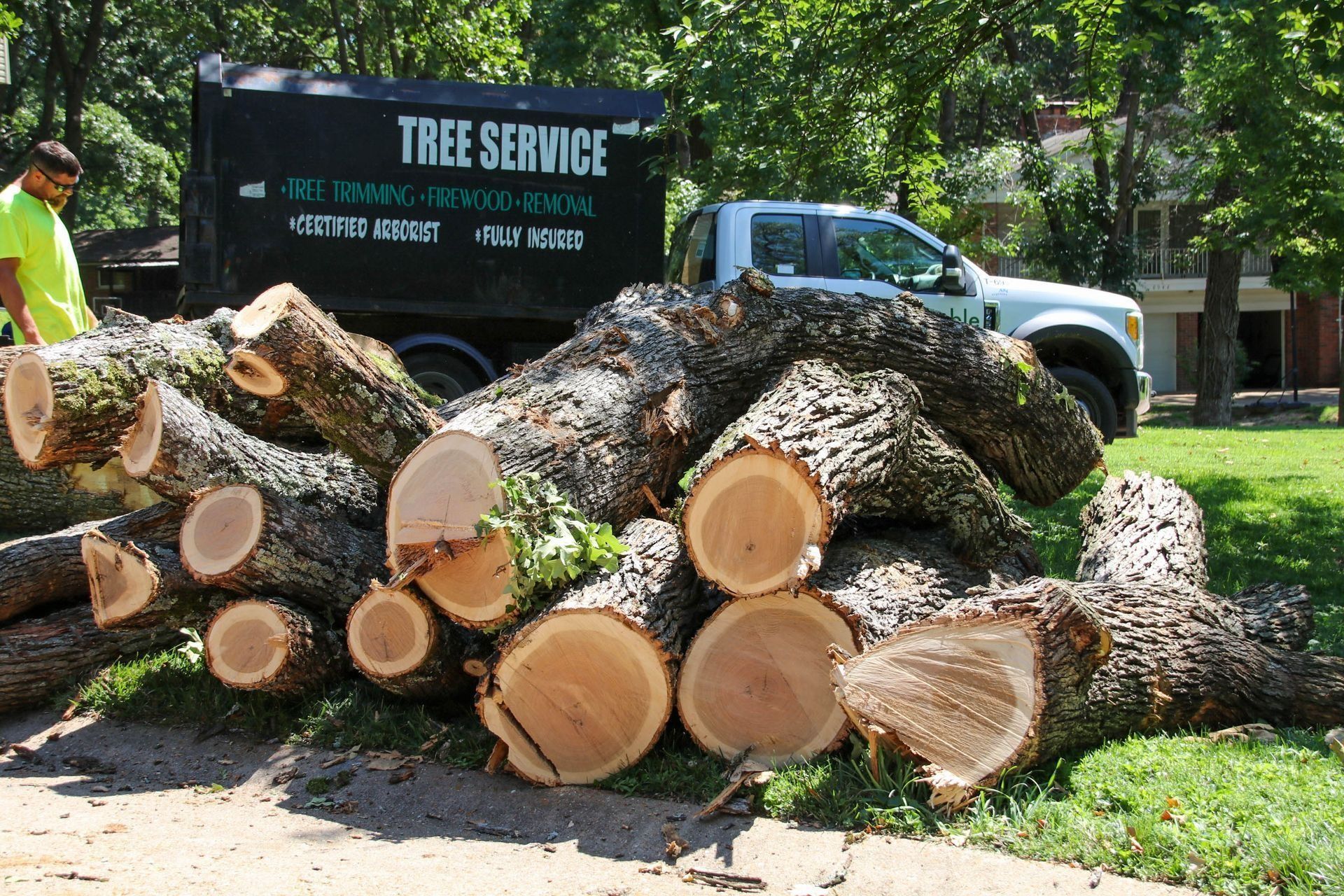 Logs of cut tree branches piled on a lawn with a tree service truck and worker in the background.