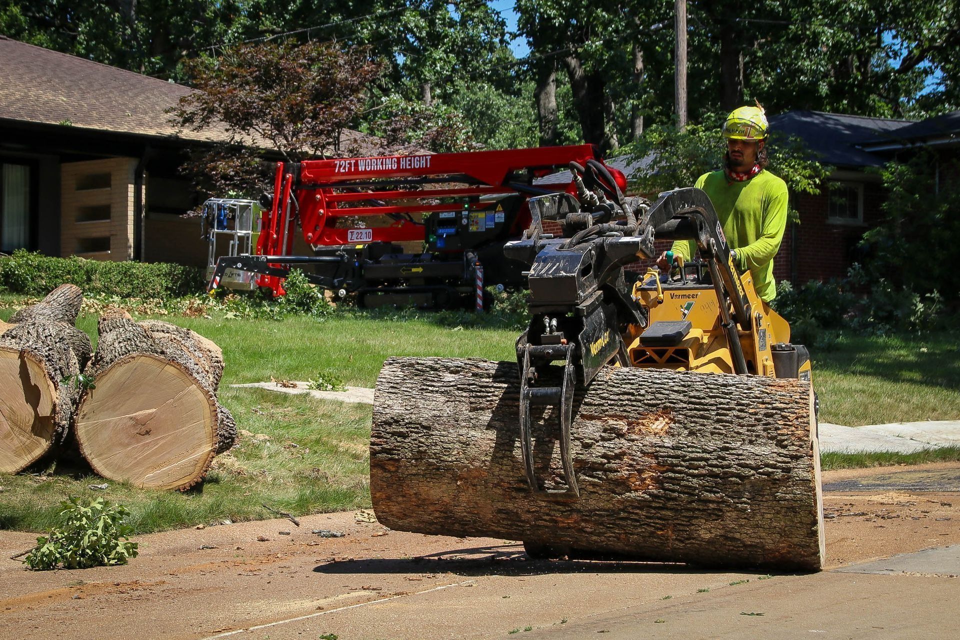 Person in yellow shirt operating a small machine lifting a large tree log on a residential street. A red truck is in the background.