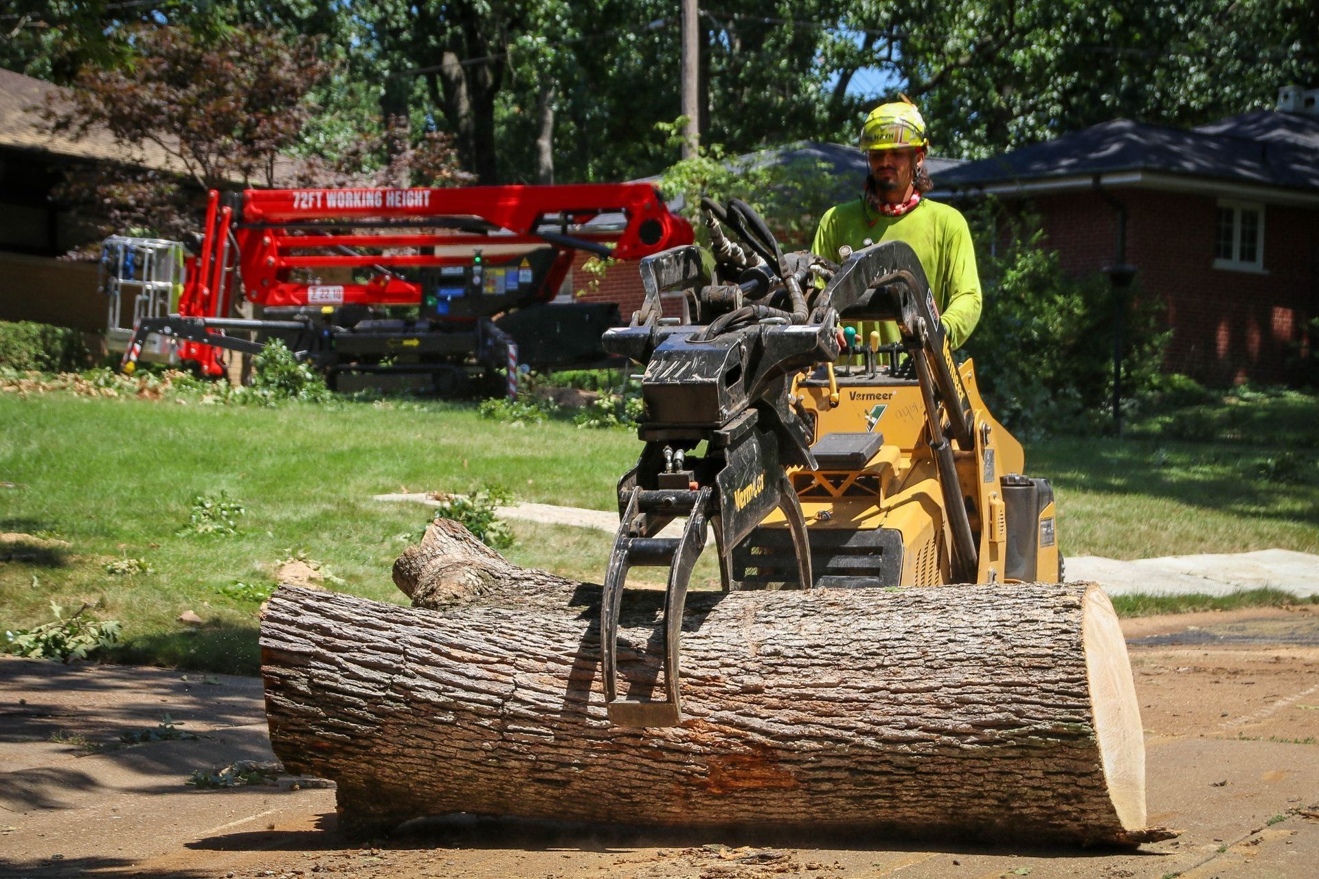 Yellow skid steer with a worker lifting a large log on a residential street. A red lift truck is in the background.