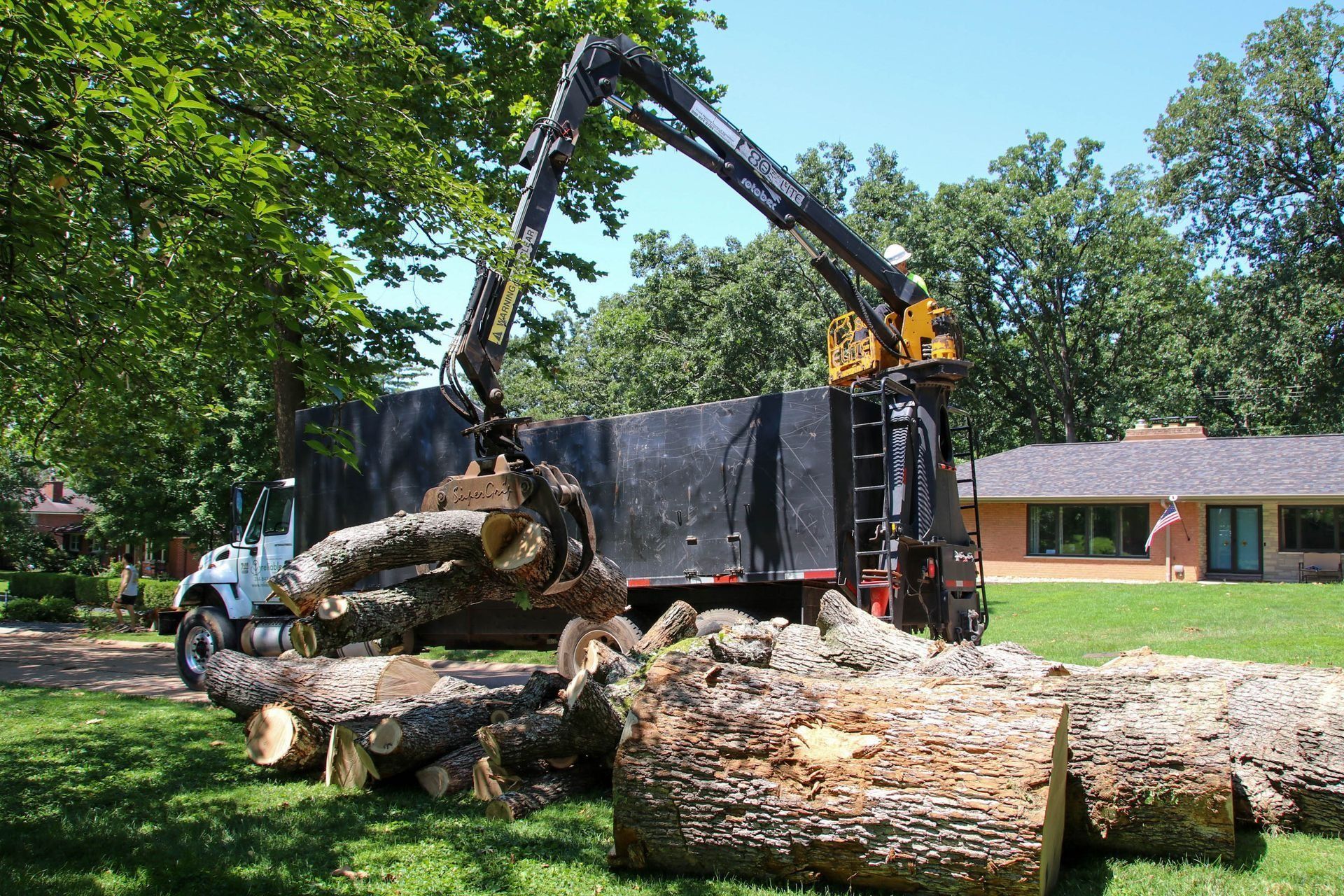 A truck with a crane loading large logs on a grassy lawn near a house.