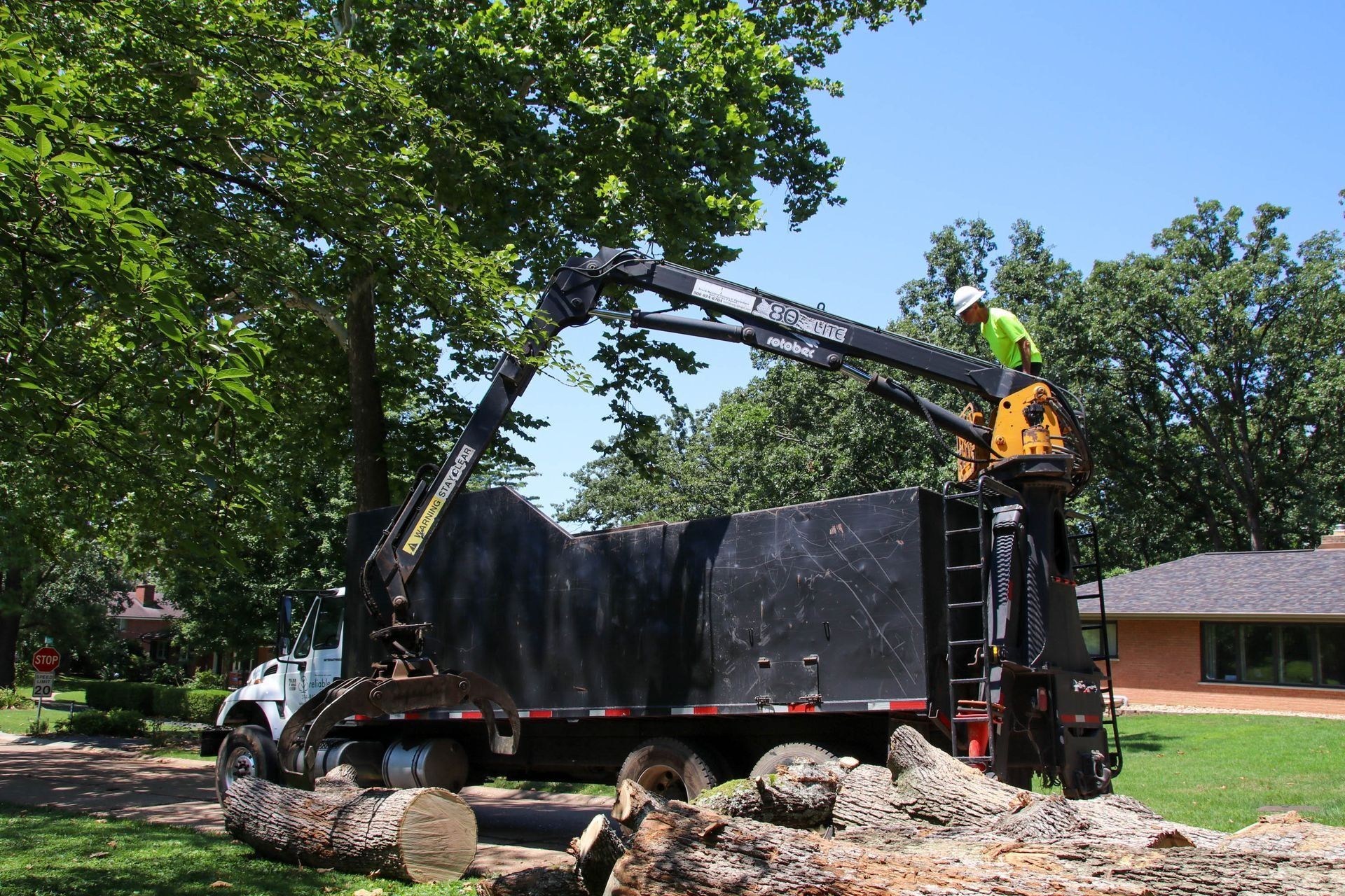 A worker loads tree logs into a truck with a mechanical arm on a sunny day.