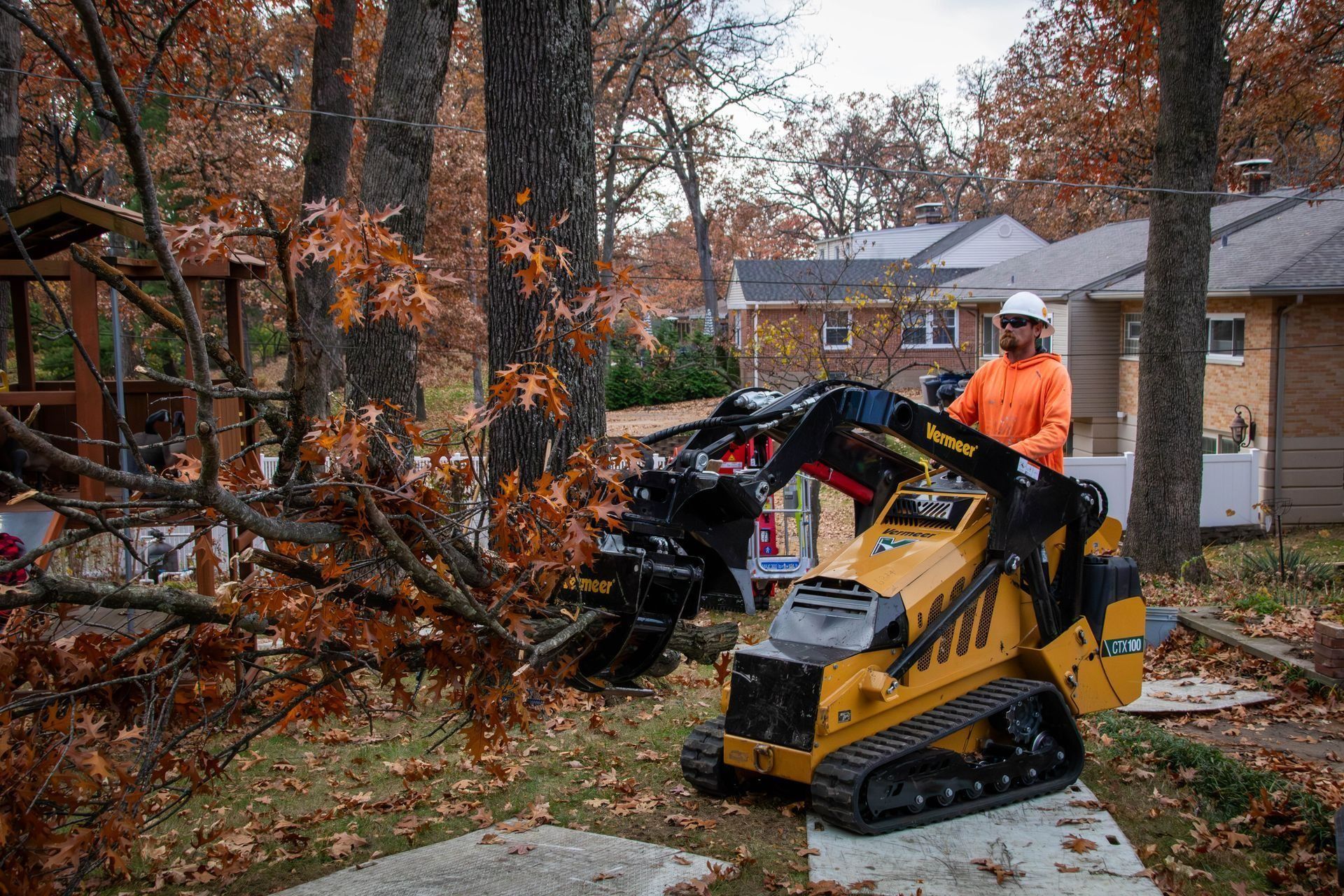 Person in orange vest operates a yellow mini excavator to cut a tree branch in a residential yard.