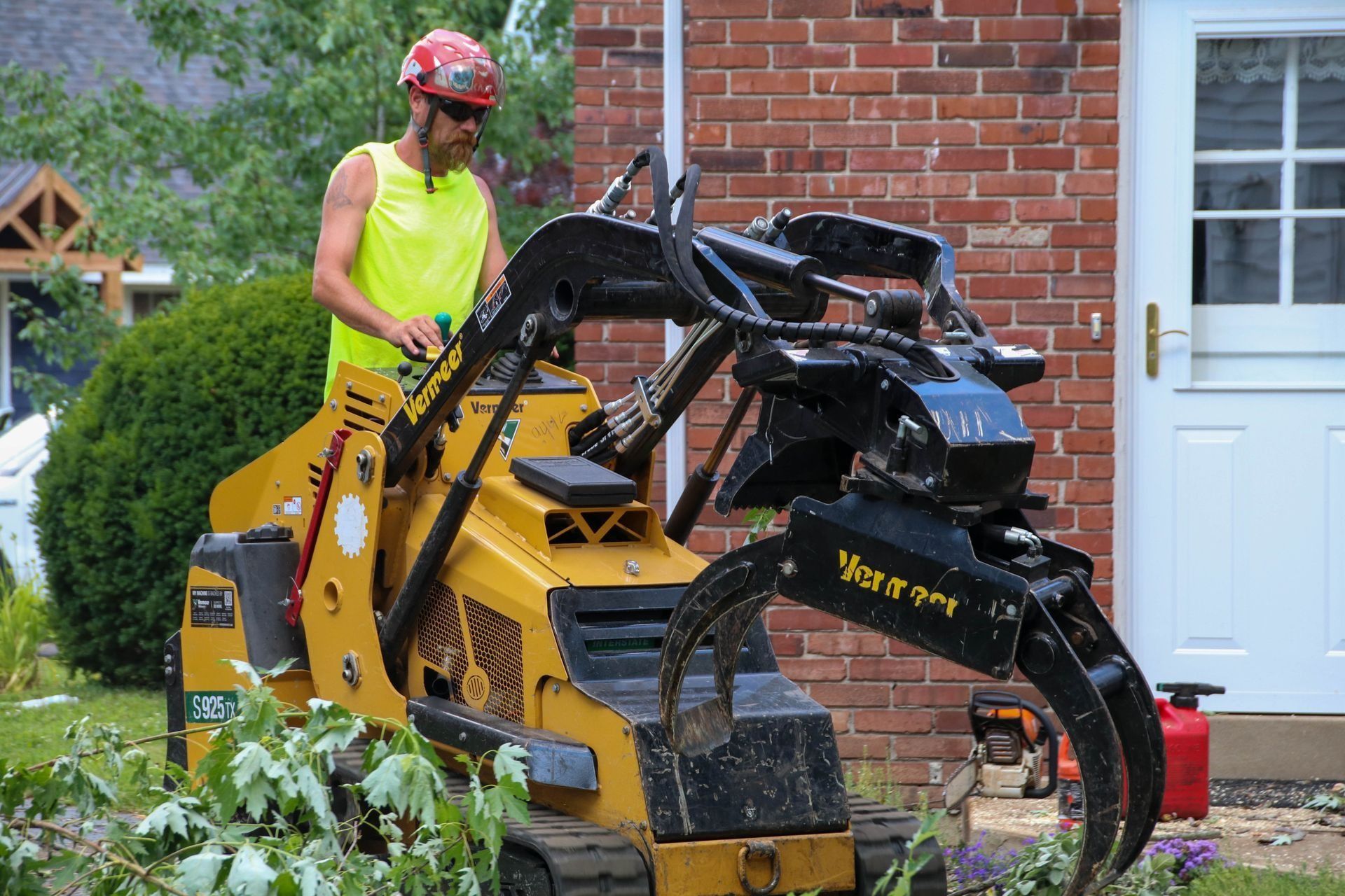 Man in a bright yellow shirt operates a yellow tree-removal machine near a brick house.