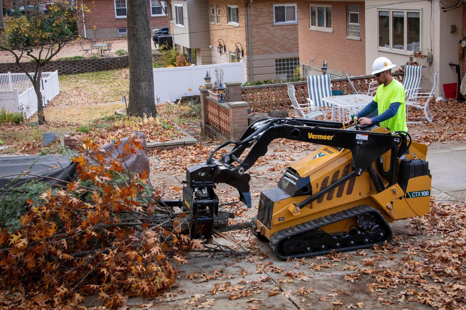 Yellow mini-excavator with a claw arm clearing branches; operator in a hardhat. Outdoor setting with fallen leaves.