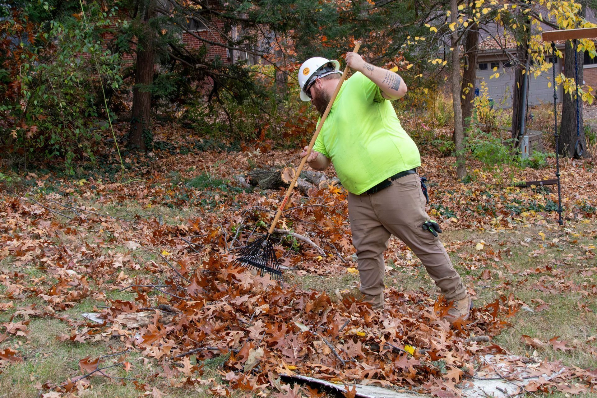 Man raking brown leaves in a yard, wearing a hard hat, neon green shirt, and tan pants.
