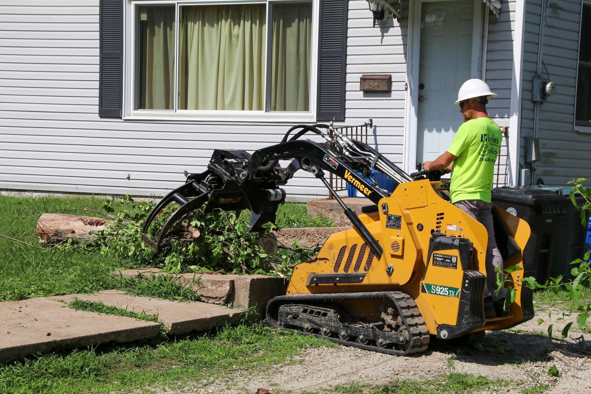 Yellow skid steer with a worker in a hard hat removes branches and debris near a house.