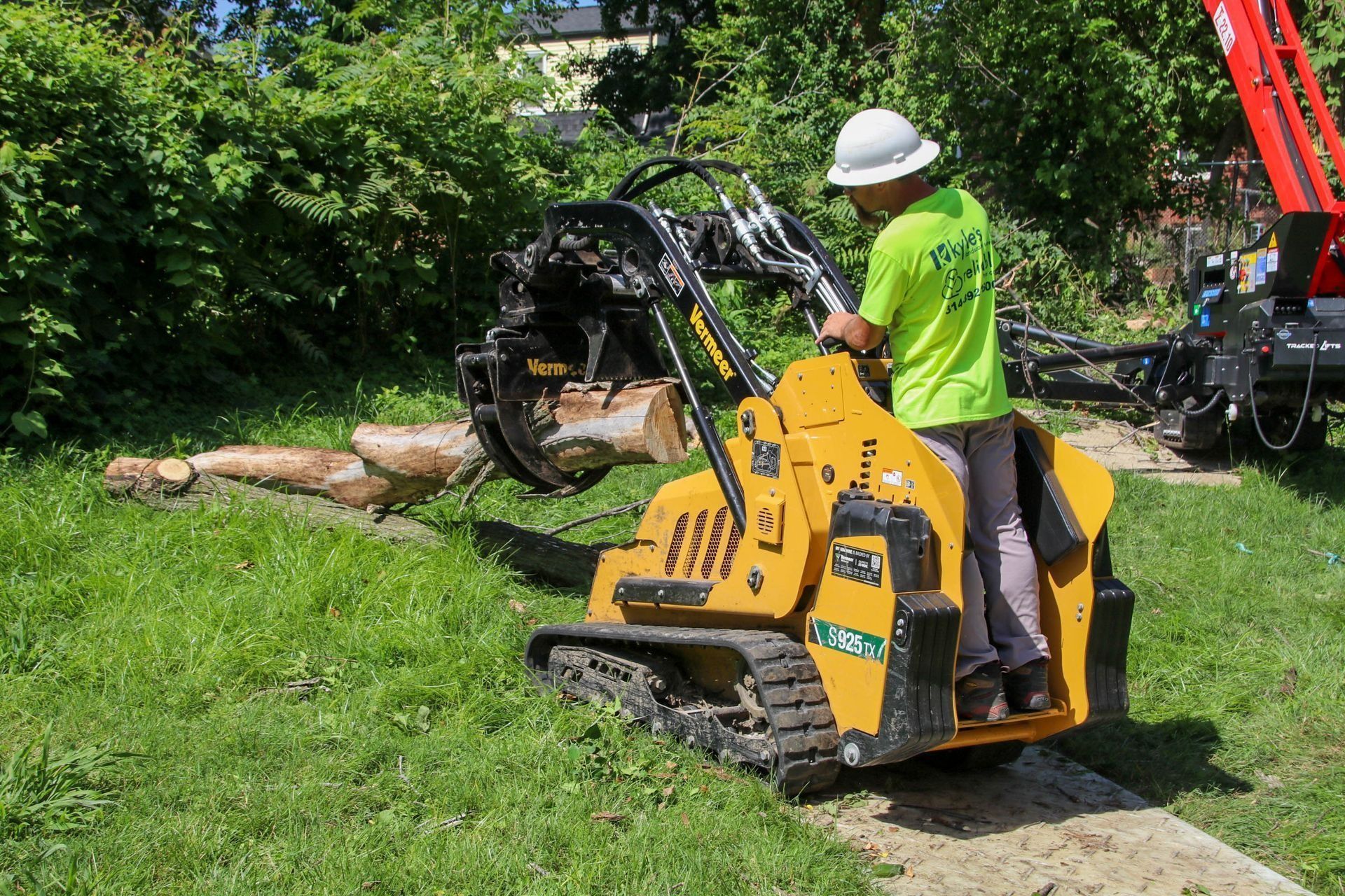 Man in neon shirt operating a tracked skid steer with a tree limb in its grapple; outdoors on grass.