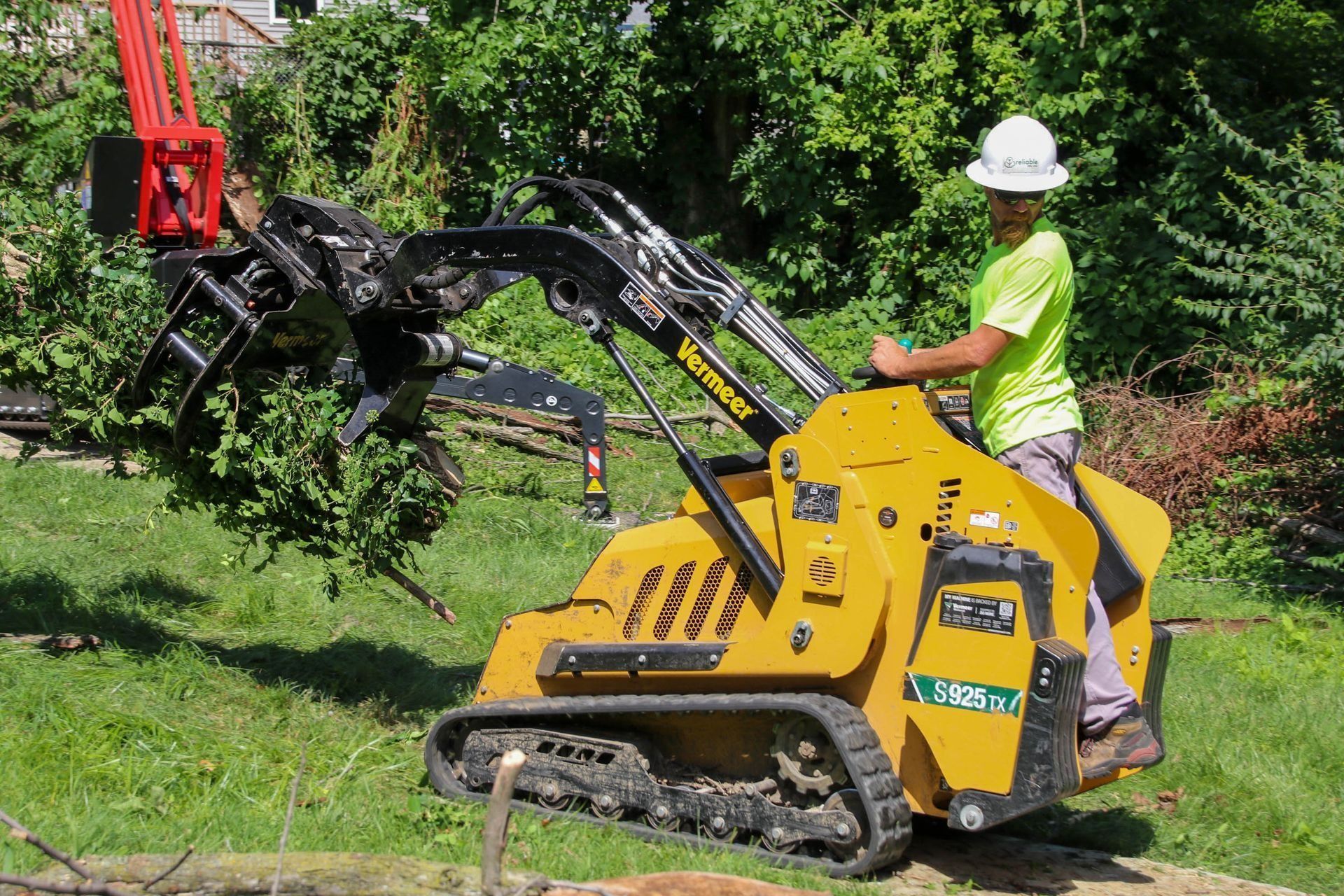 Person operating a yellow mini-skid steer with tree shears, cutting branches in a grassy yard.