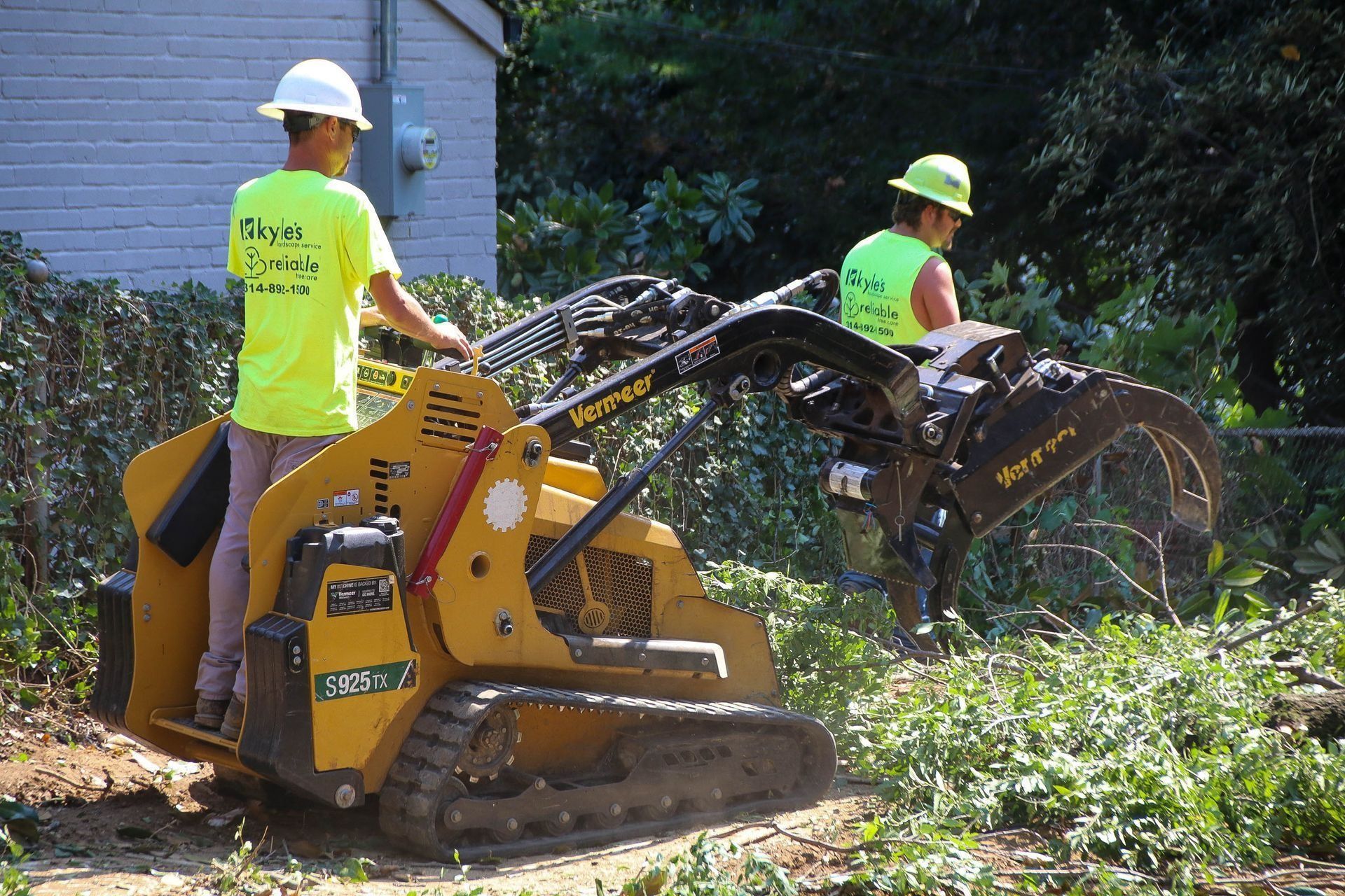 Two workers using a yellow skid steer with a tree shear to clear brush near a building.