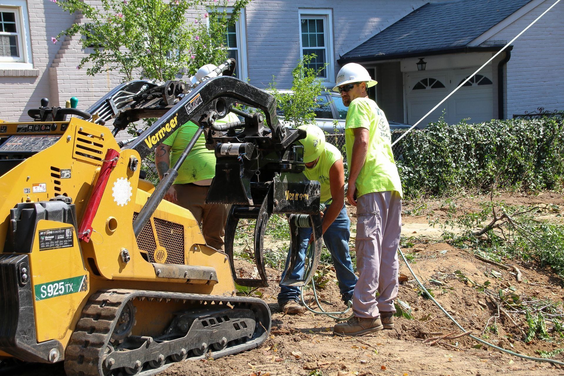 Workers in safety vests operating machinery near a house; one is looking at the equipment.