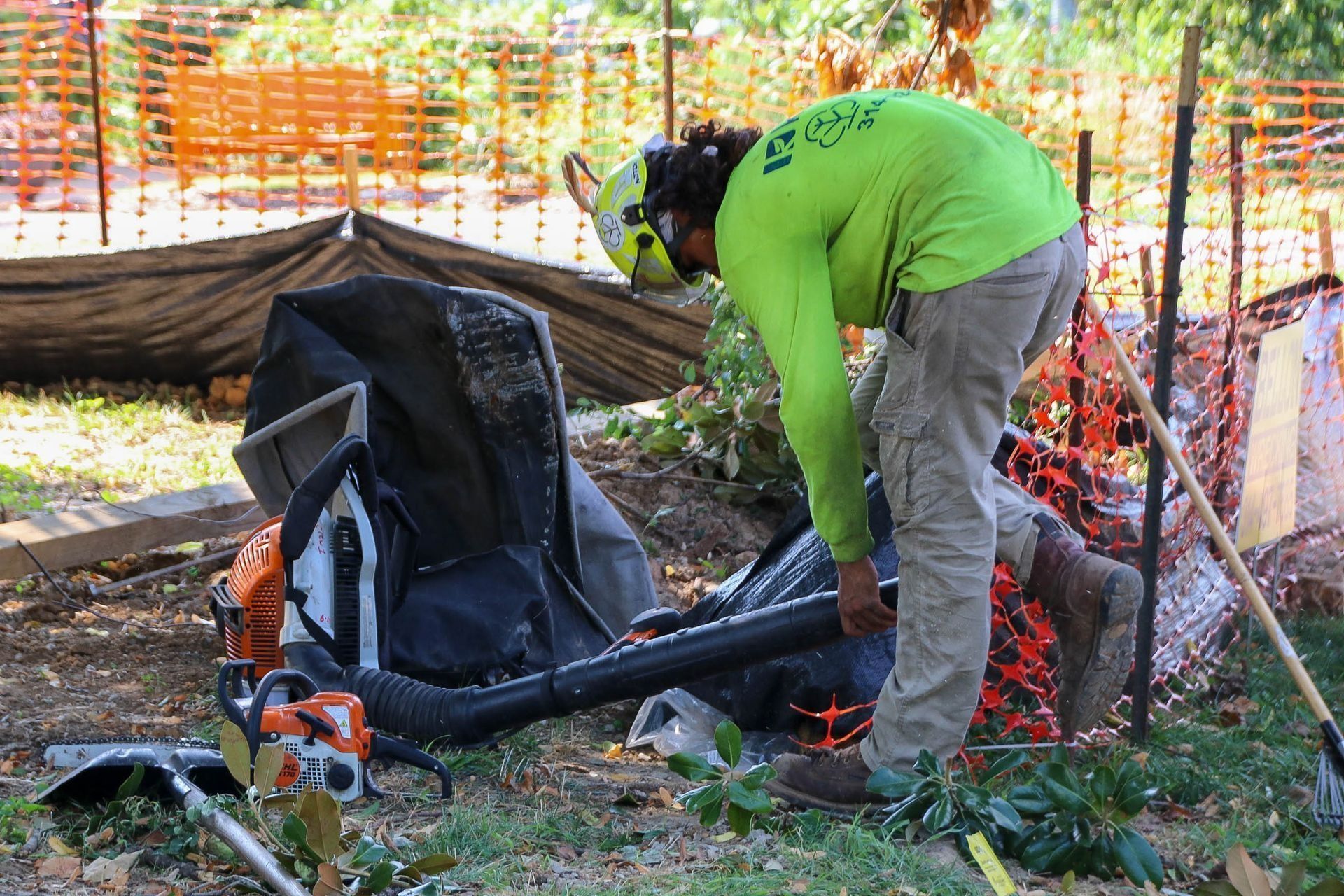 Worker in safety gear uses a leaf blower outdoors, near a chainsaw and fencing.