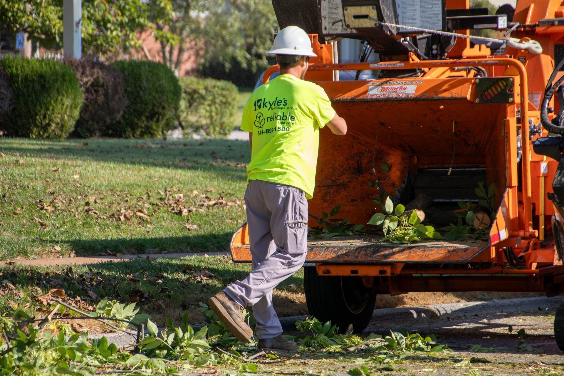Person in a neon yellow shirt feeding green branches into an orange wood chipper on a sunny day.