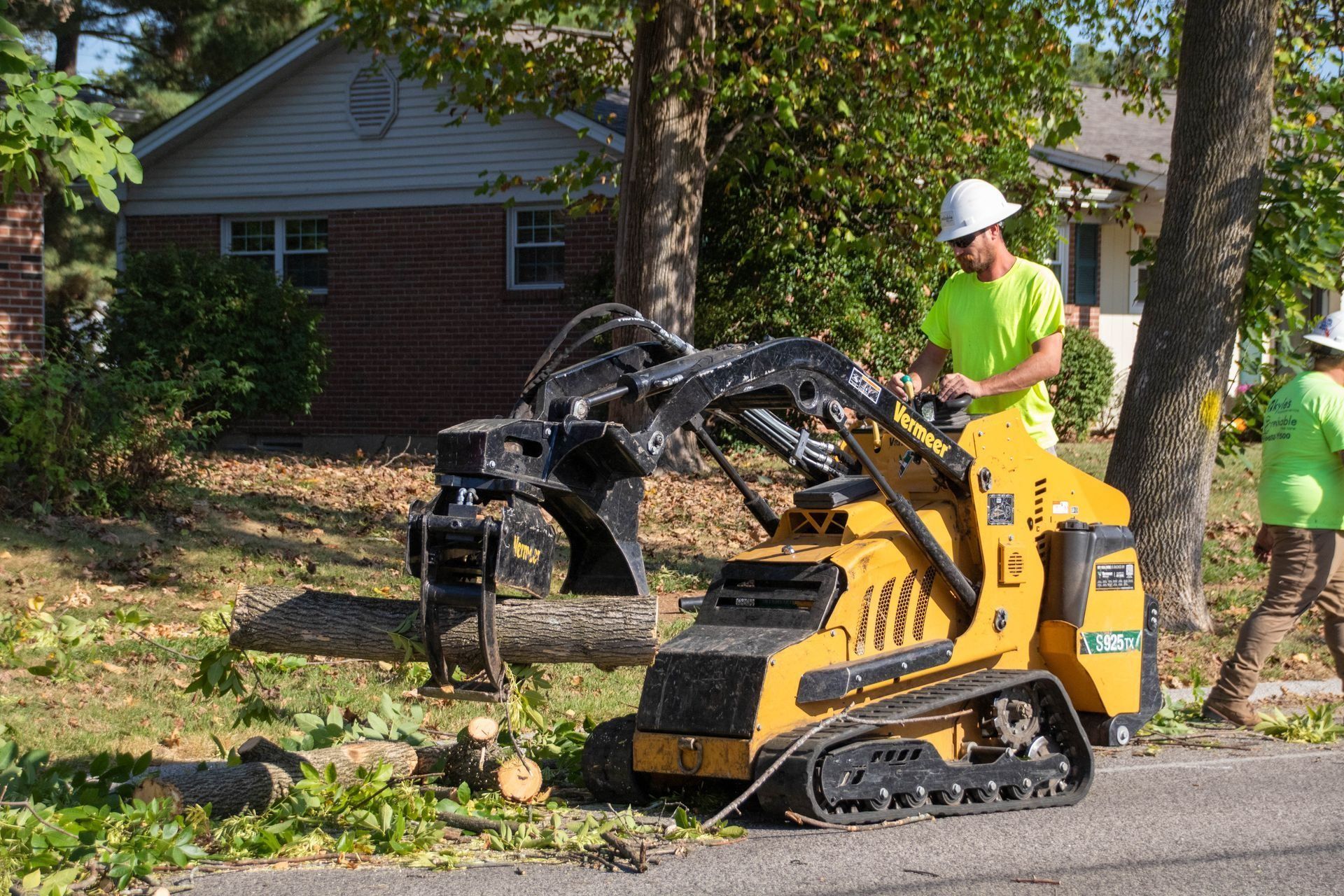 Two workers use a tracked mini-excavator to remove a tree branch on a residential street.