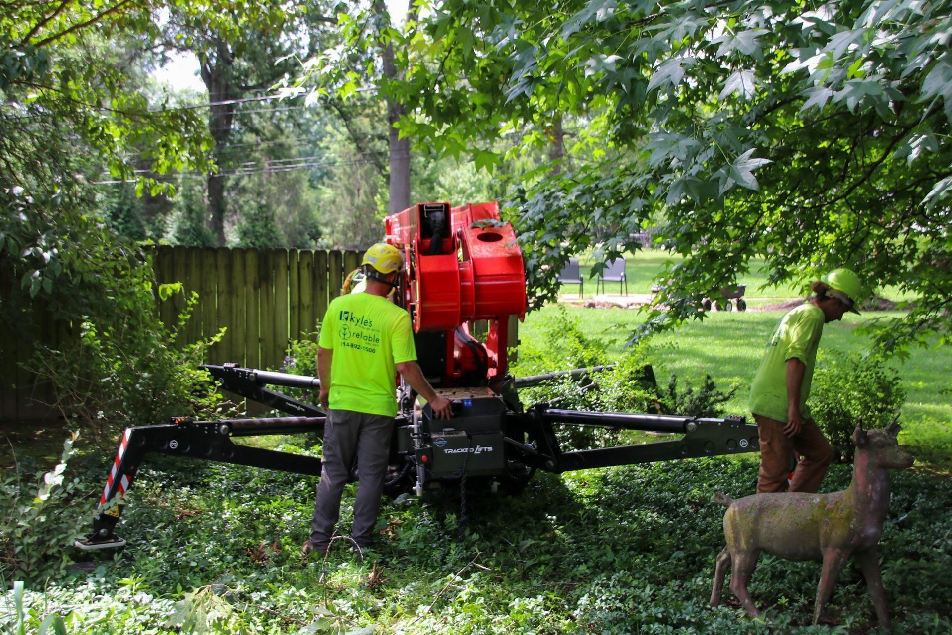 Two workers operating a red tree-chipping machine in a backyard. One is wearing a green shirt, and the other is near a deer statue.