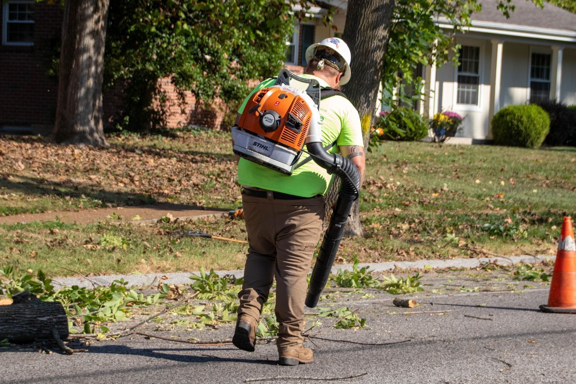 A person wearing safety gear uses a leaf blower on a road, blowing debris.
