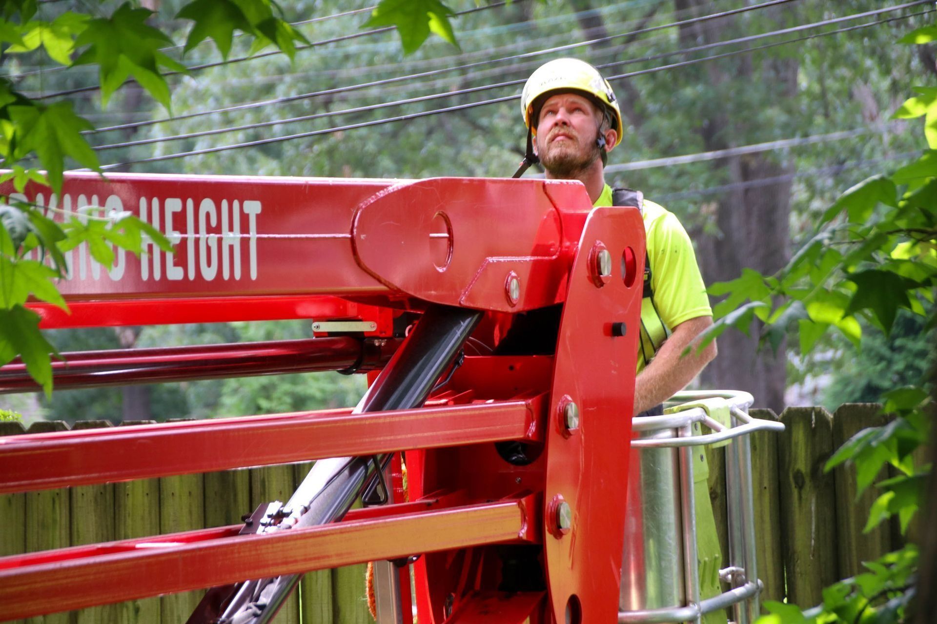 Worker in a yellow shirt and hard hat operates a red boom lift, looking upwards.