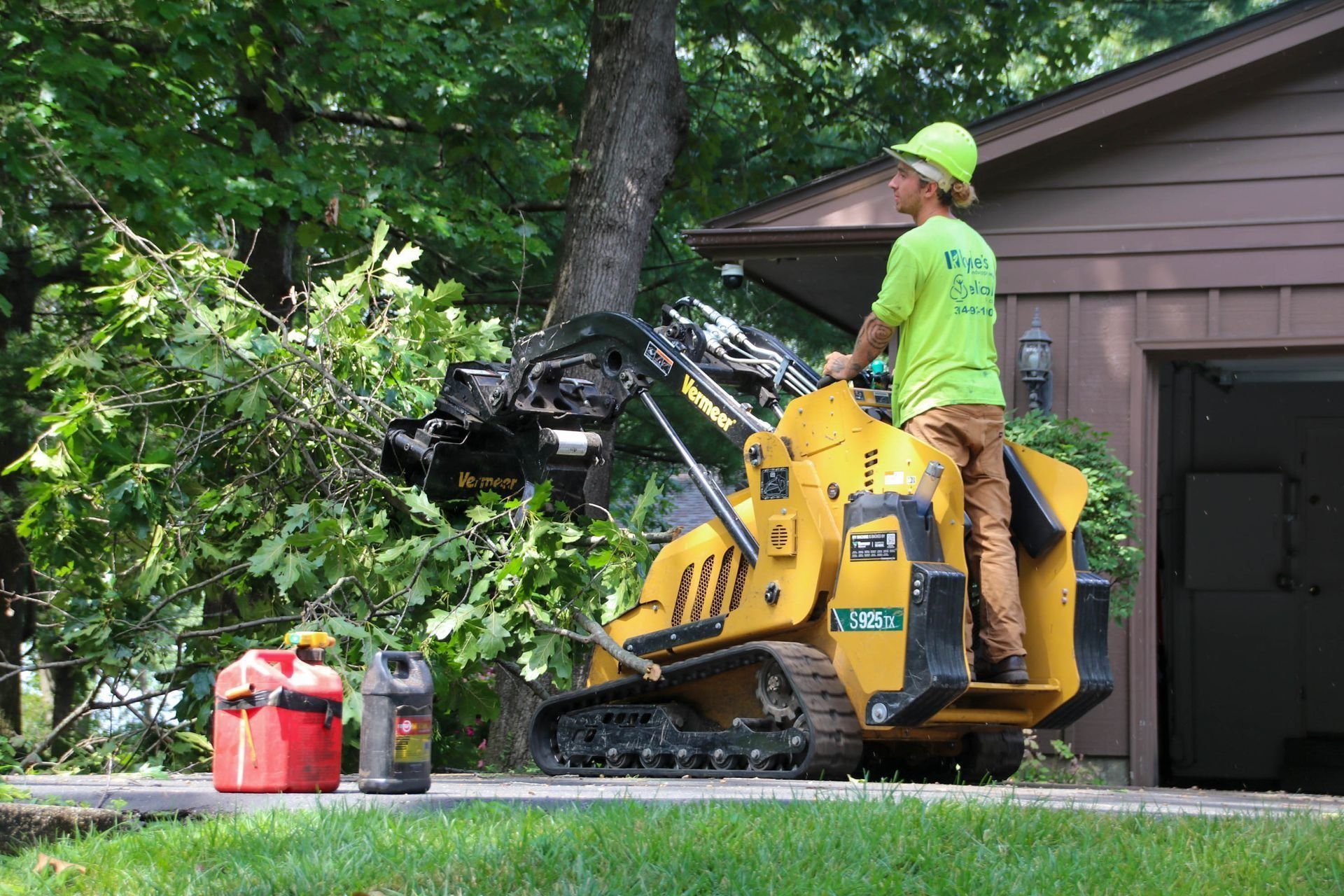 A person in a hard hat operates a yellow skid steer, lifting tree branches in front of a garage.