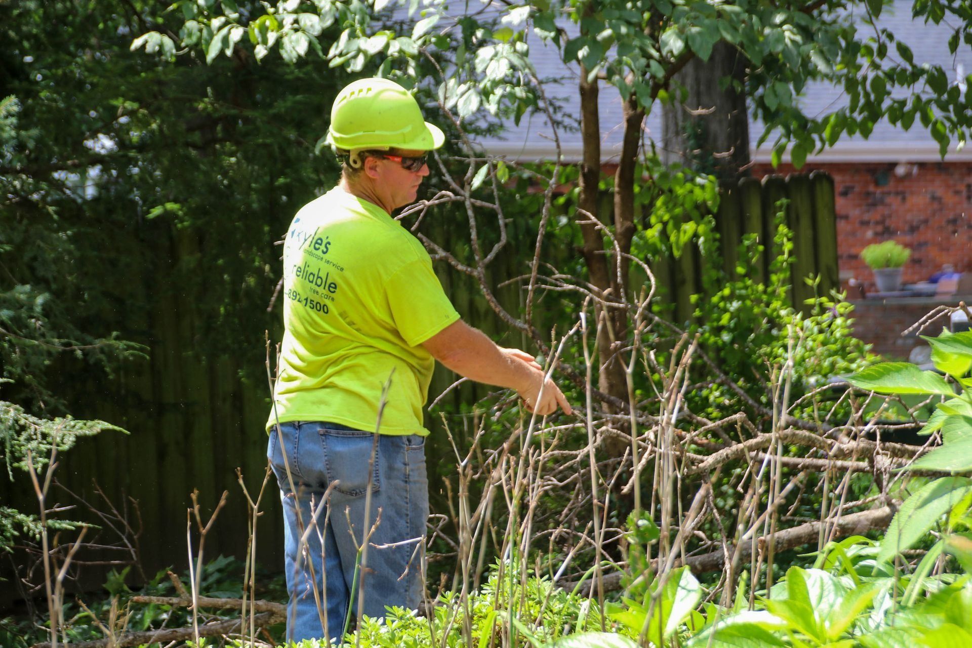 Person in a yellow hard hat and shirt trimming dry branches in a sunny garden.