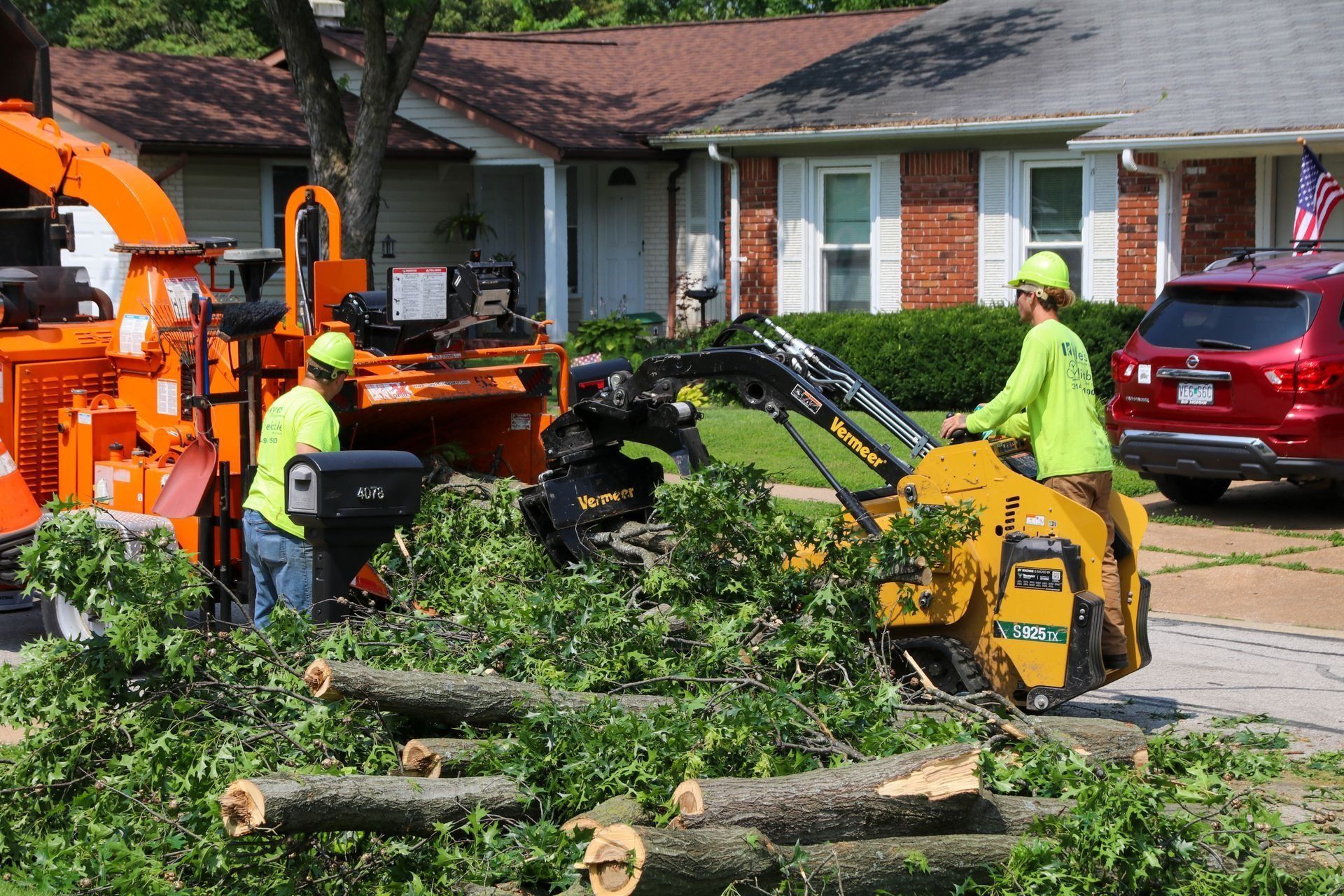 Two tree service workers in safety vests operating equipment to clear branches on a residential street.
