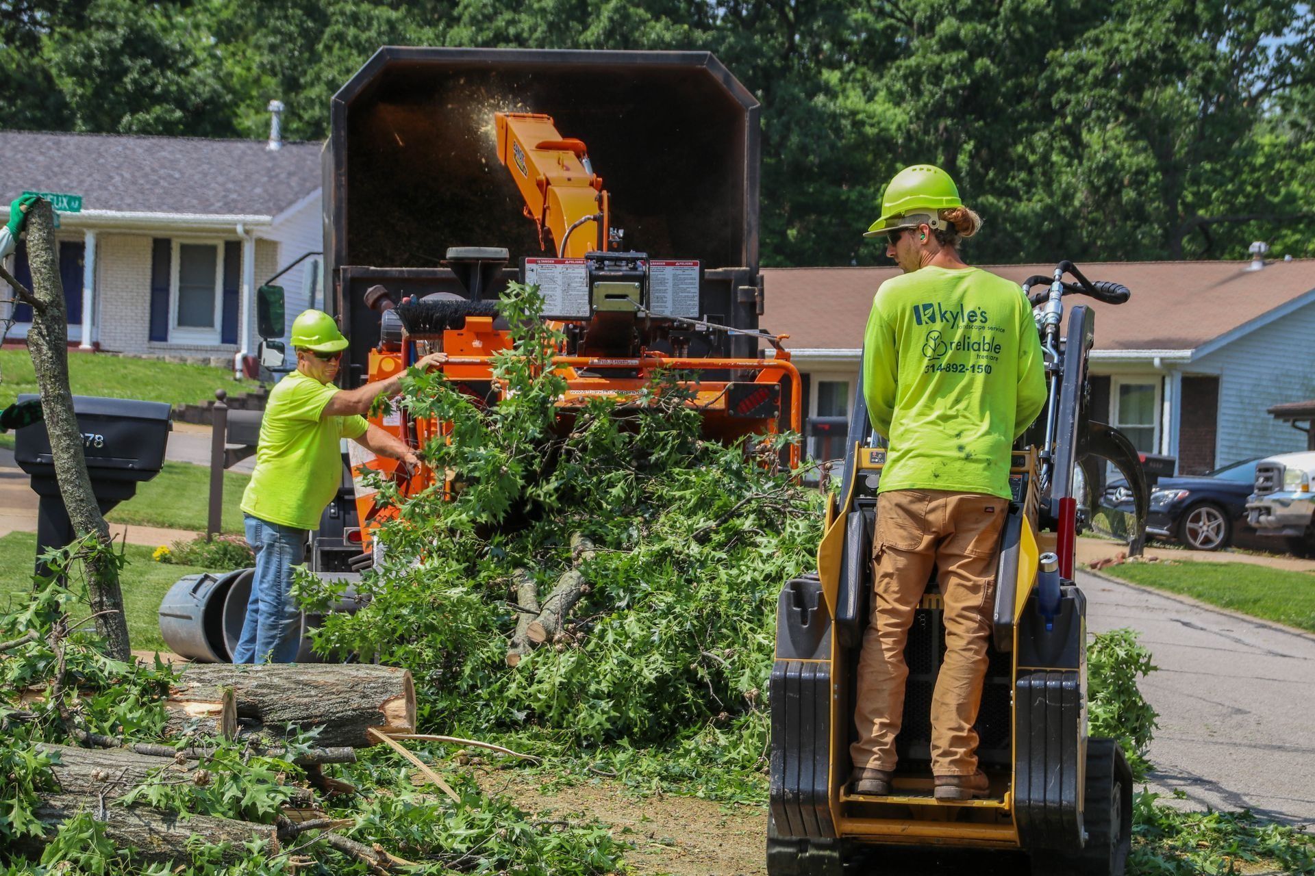 Two workers in green shirts feed branches into a wood chipper in a residential area.