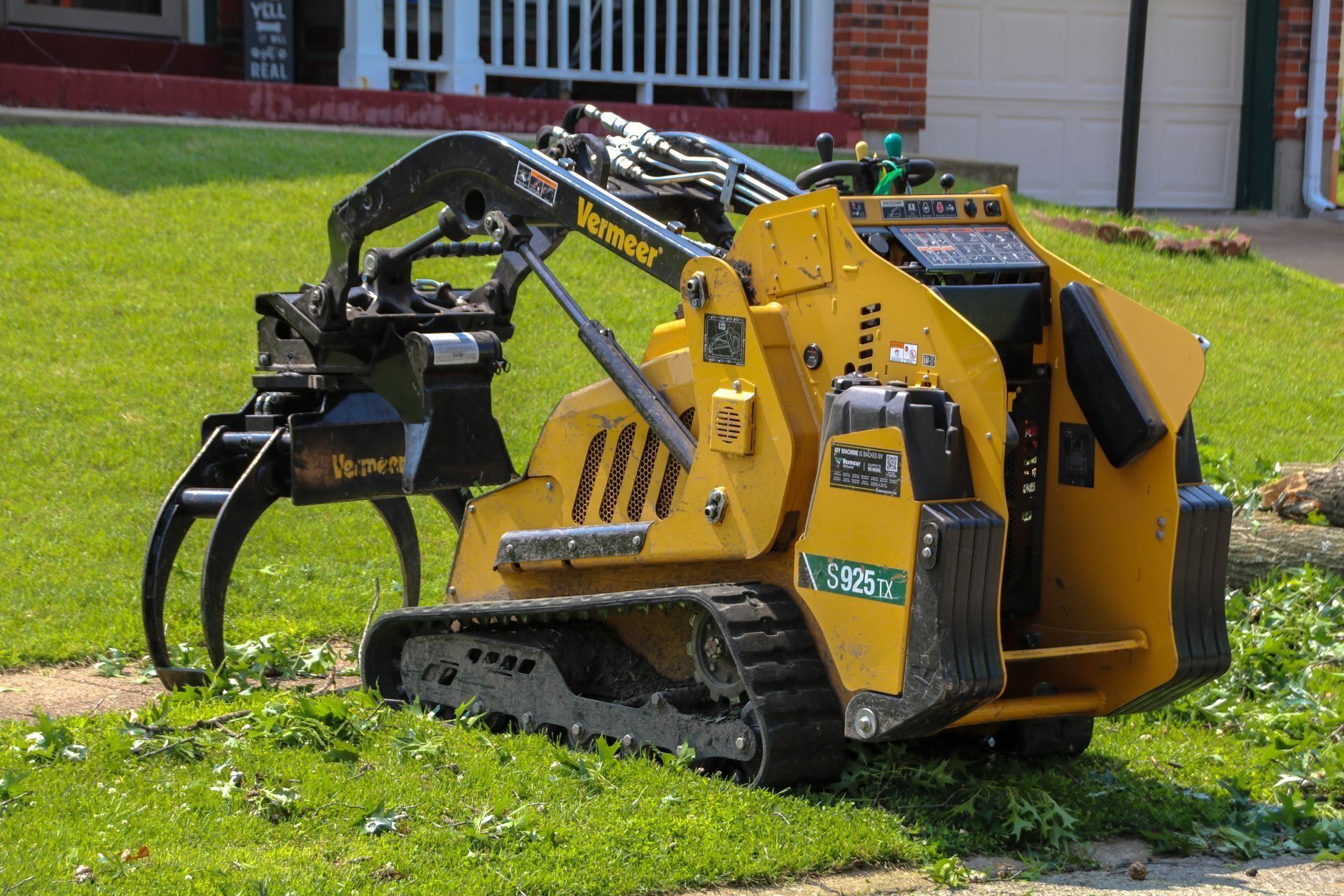Yellow mini-skid steer with tree shear attachment on grass near a house.