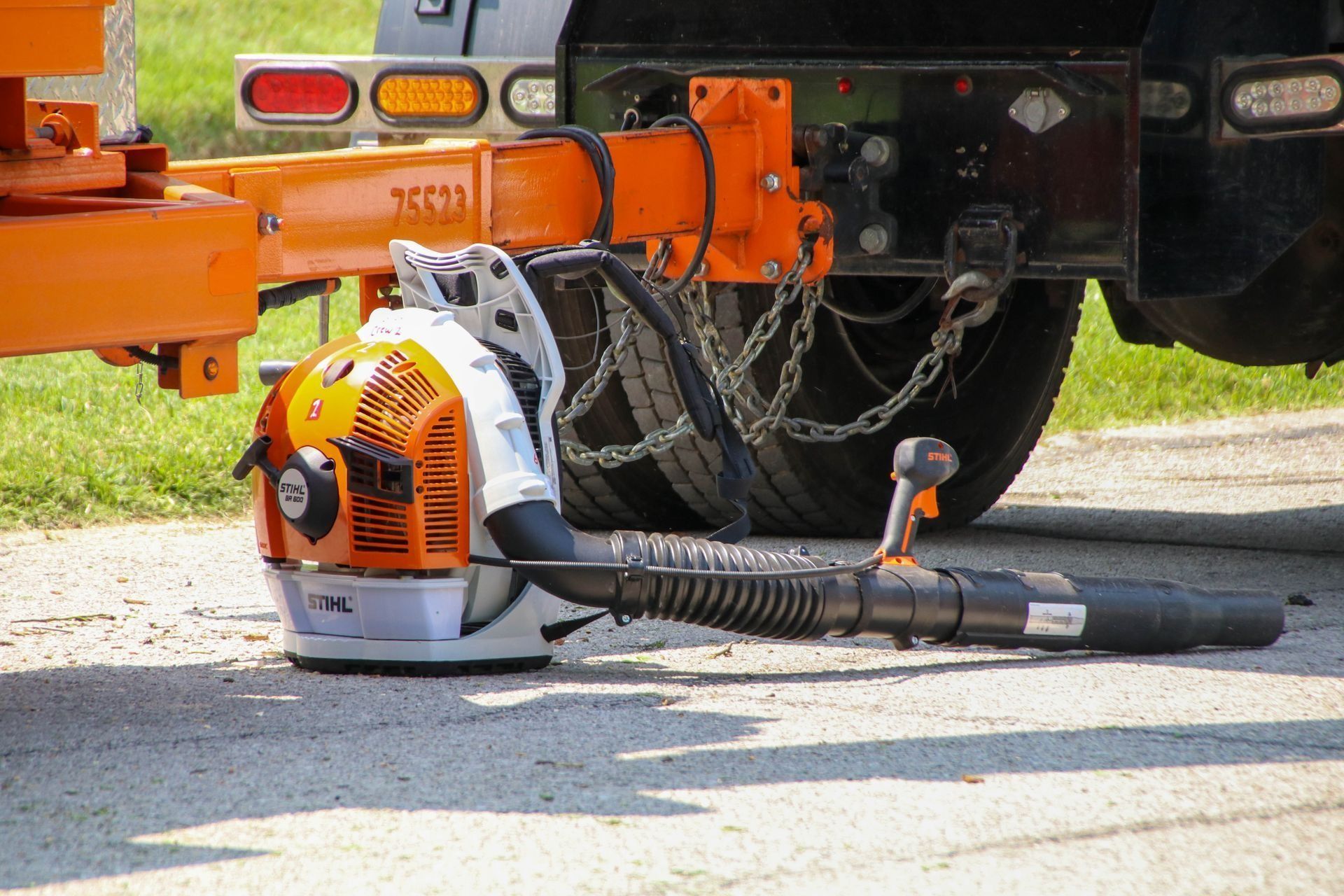 An orange and white leaf blower rests on pavement near an orange truck hitch.