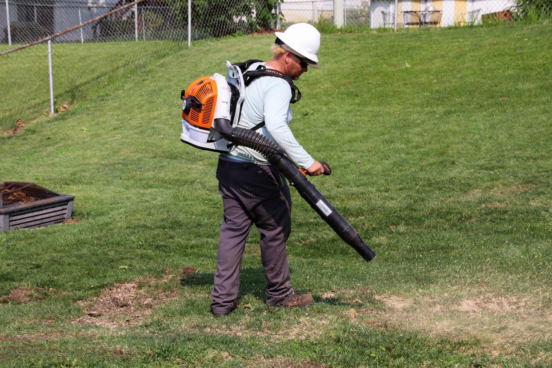 Person with a backpack blower, wearing a hard hat, blowing grass on a lawn.