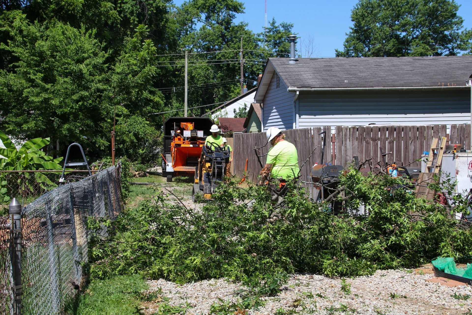 Two people in safety vests chipping branches near a residential fence and house.