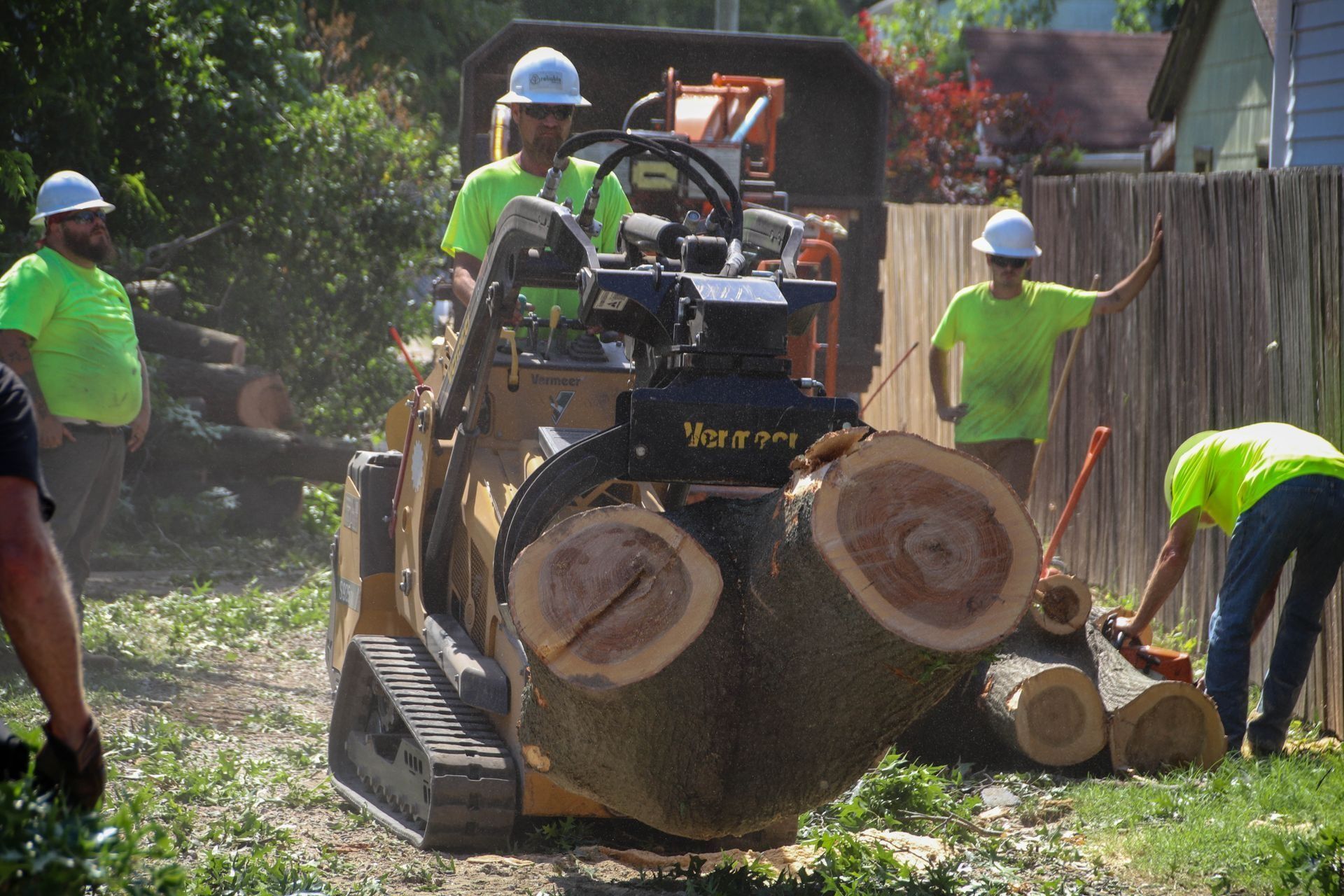 Workers in lime green shirts and hard hats use a skid steer to move tree logs near a fence.