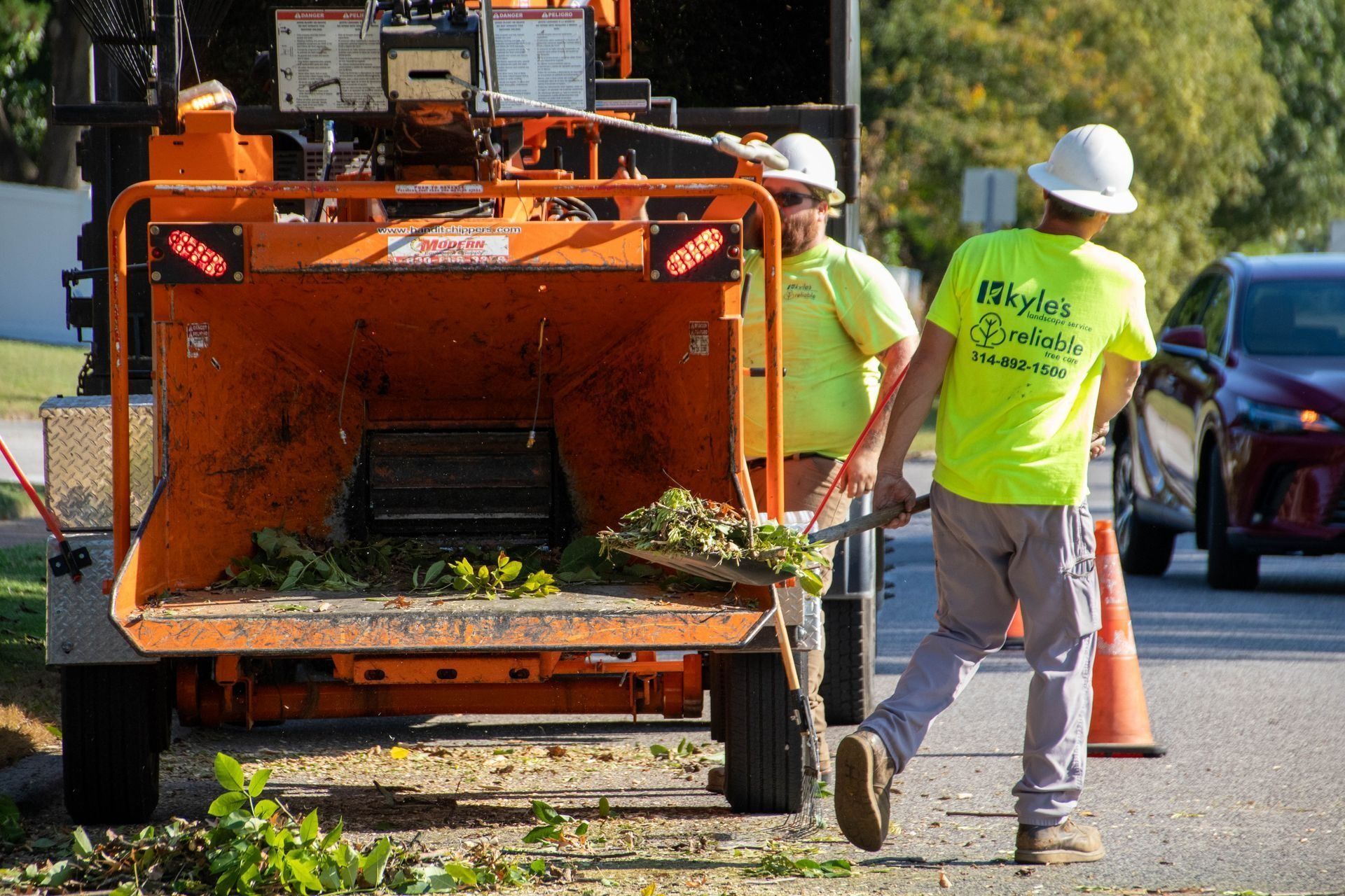 Two workers feeding yard waste into an orange wood chipper on a street.