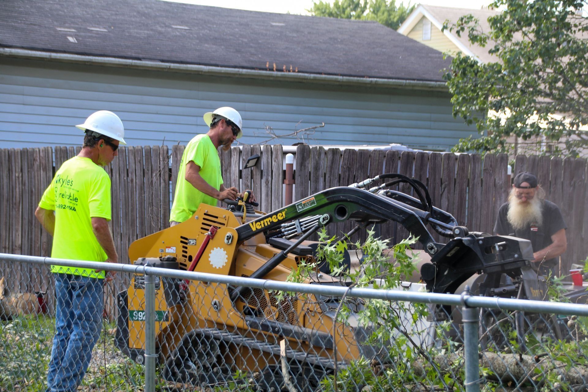 Two workers using a stump grinder on overgrown brush next to a fence. One man watches.