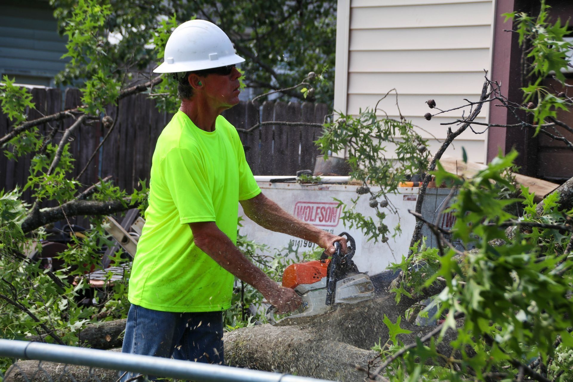 Man in white hard hat and neon shirt using chainsaw on fallen tree branches.