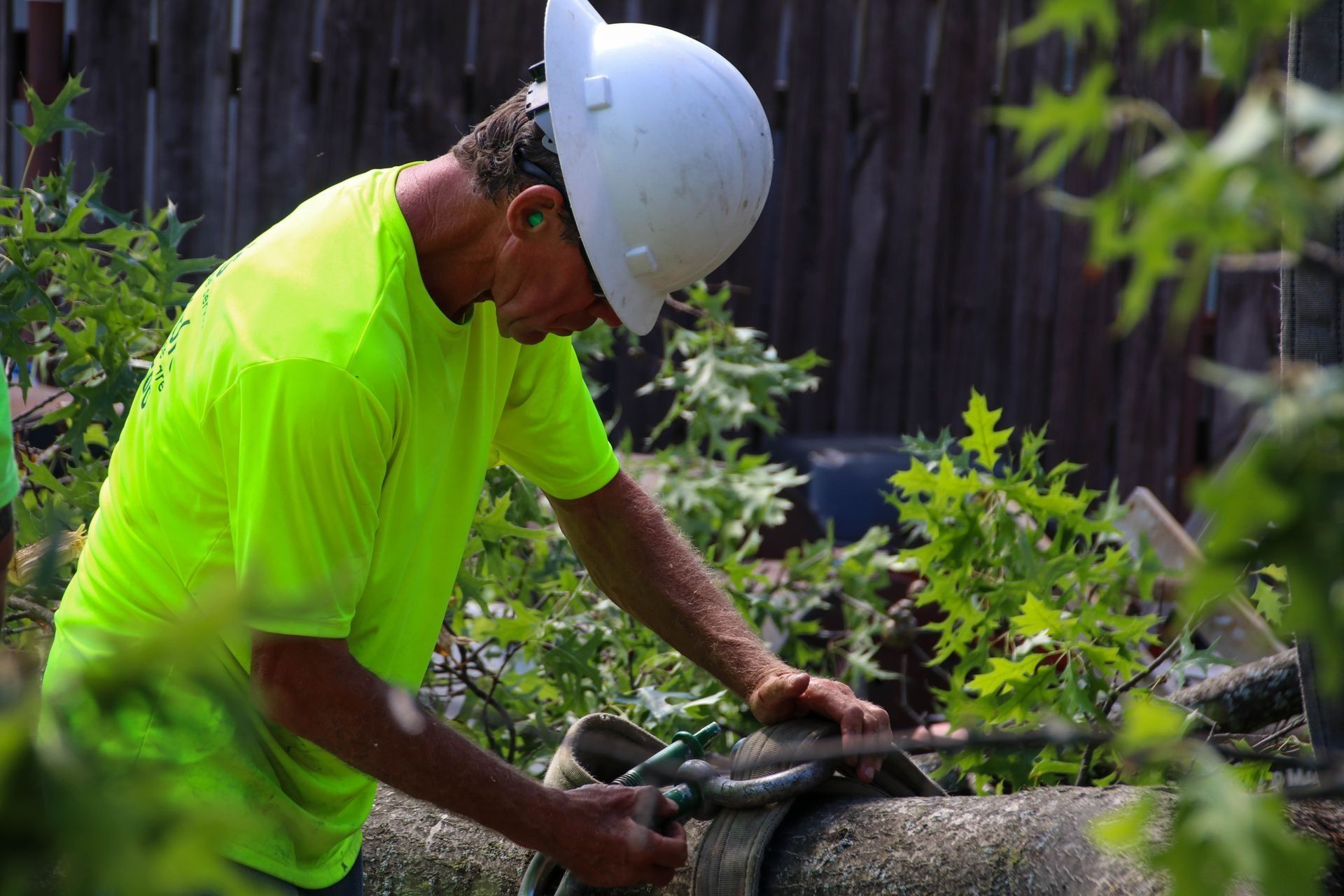 Man in neon shirt and hard hat working on log, surrounded by branches.