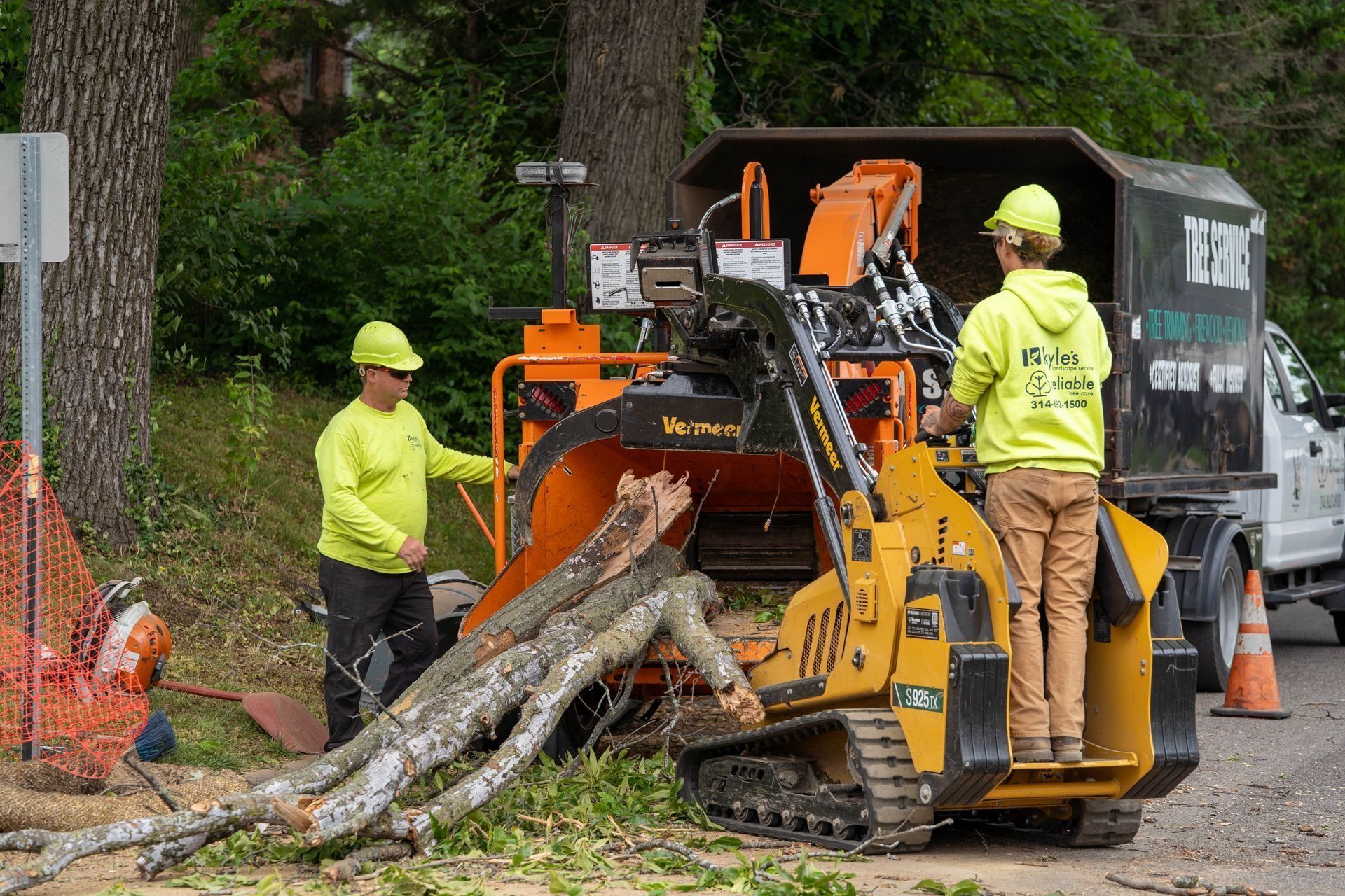 Two workers in neon yellow shirts operating a wood chipper to process tree branches next to a truck.