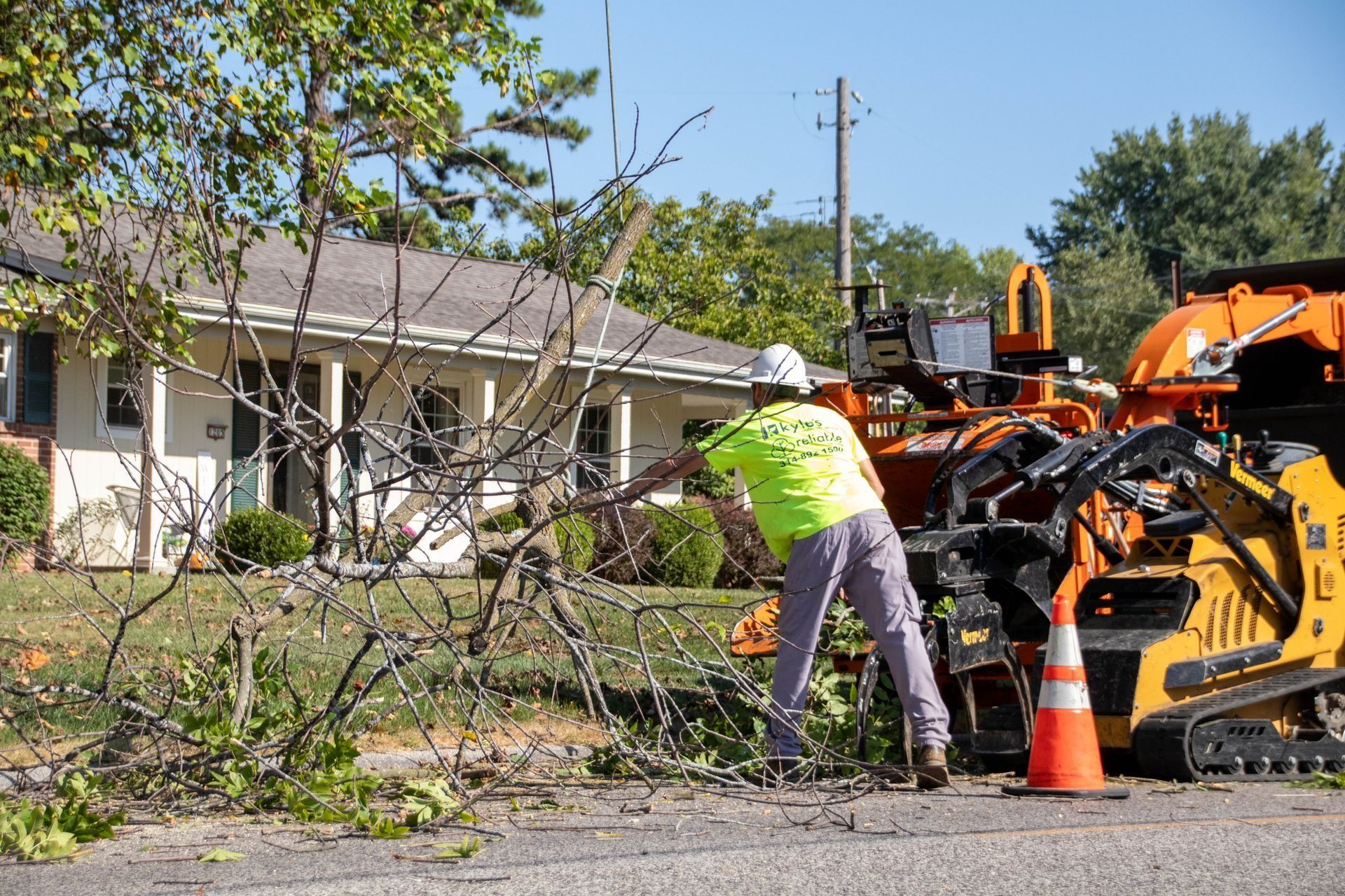 Arborist in a lime green shirt feeds tree branches into a wood chipper near a house.