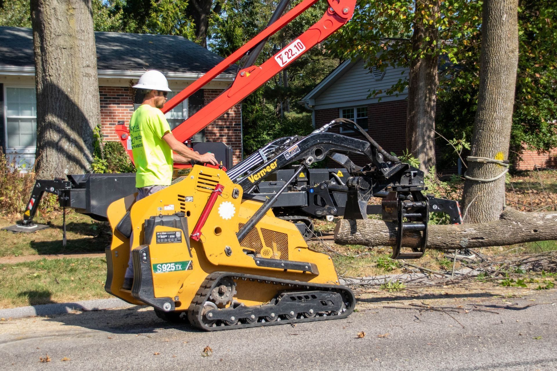 Man in hard hat operating a small yellow tracked machine with an arm and grapple, cutting a tree trunk.