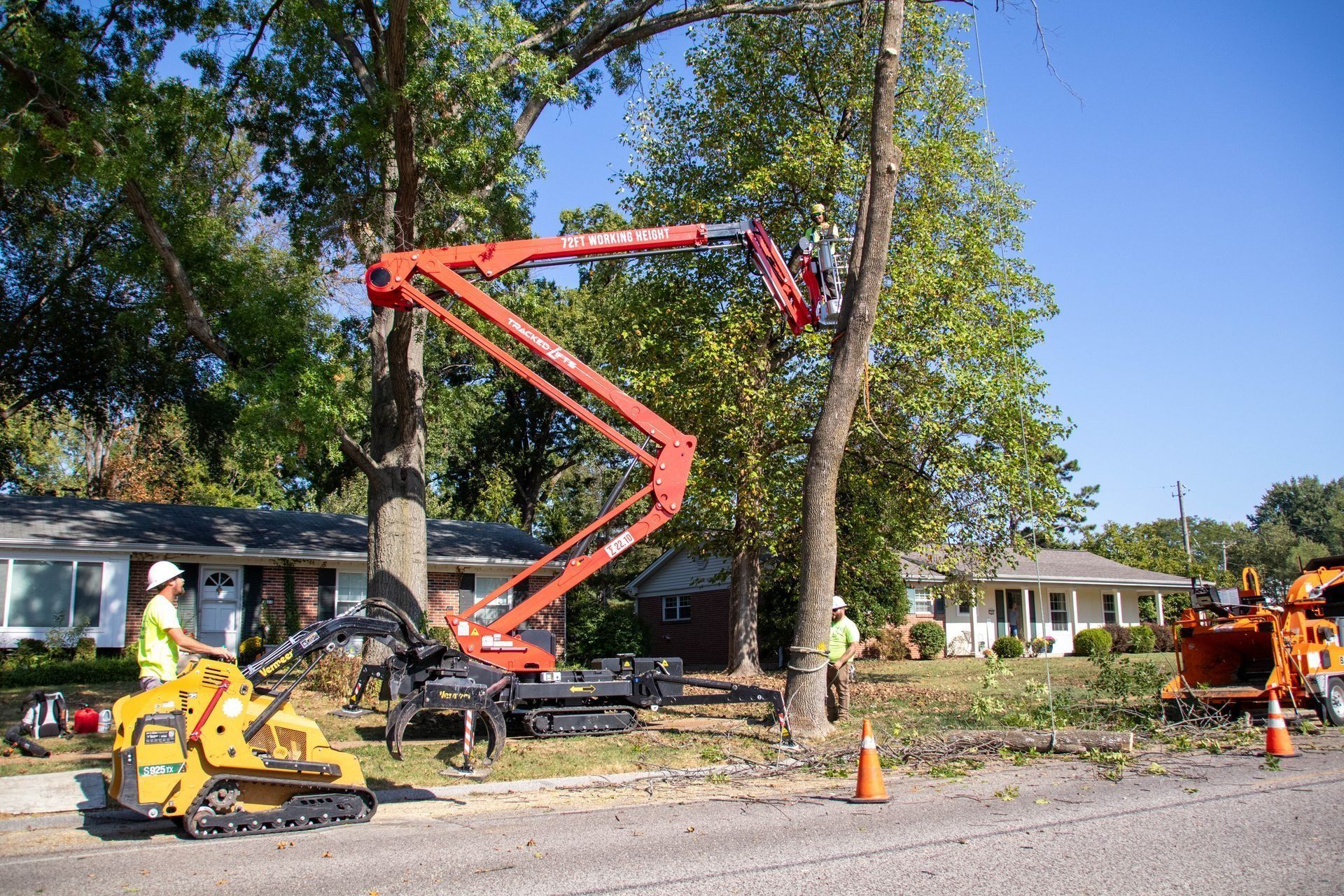 Tree trimming service in progress; cherry picker, chipper, workers on residential street.