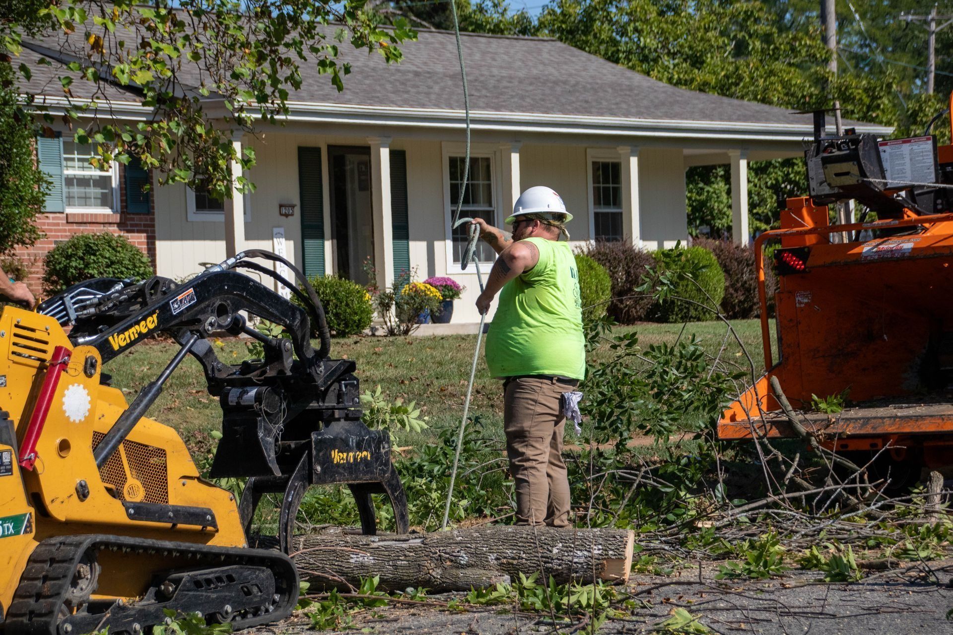 Tree service worker near a house, operating equipment. Orange and yellow machinery, green shirt, white hard hat.