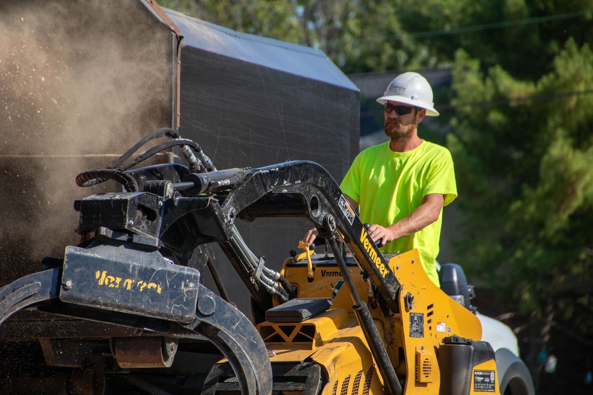 Man operating a yellow skid steer, wearing a hard hat and neon shirt, with dust billowing as he works.